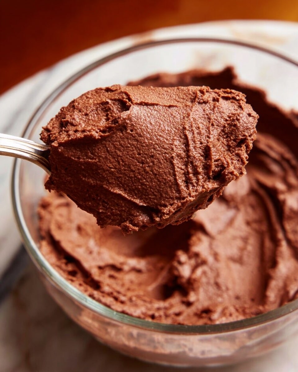 A close-up view of a glass bowl filled with rich, thick, dark brown chocolate mousse. The mousse has a smooth but textured surface with small ridges and swirls, showing its creamy and fluffy nature. A silver spoon holds a large scoop of the mousse above the bowl, highlighting the dense and soft texture. The background features a white marbled surface under warm lighting. photo taken with an iphone --ar 4:5 --v 7