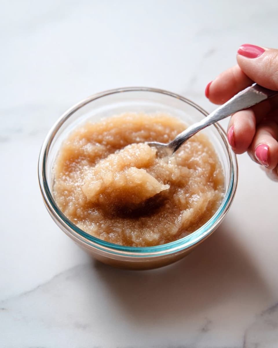 A clear glass bowl filled with a light brown chunky puree with a soft, moist texture. A woman's hand is holding a small silver spoon, dipping it into the puree near the edge of the bowl. The bowl is placed on a white marbled surface with soft natural light illuminating the scene. photo taken with an iphone --ar 4:5 --v 7