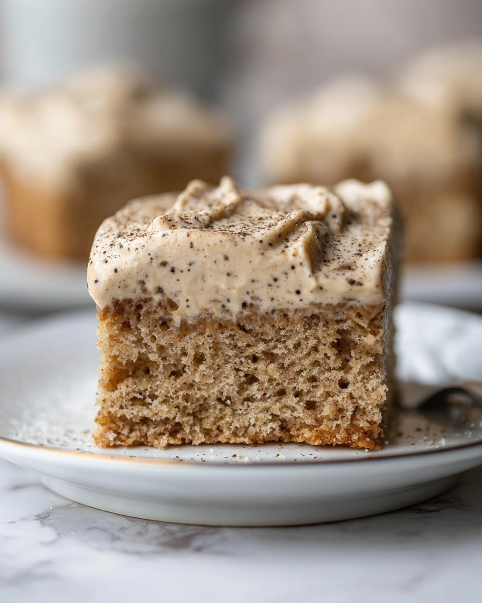 A square piece of cake sits on a white plate with a thin gold rim, placed on a white marbled surface. The cake has two layers: the bottom layer is light brown and slightly textured like sponge, while the top layer is thick, creamy, and light tan with small ridges visible on its surface. The frosting is sprinkled with white cylindrical sprinkles and a light dusting of brown powder. A silver fork with a shiny finish lies next to the cake on the plate. Photo taken with an iphone --ar 4:5 --v 7