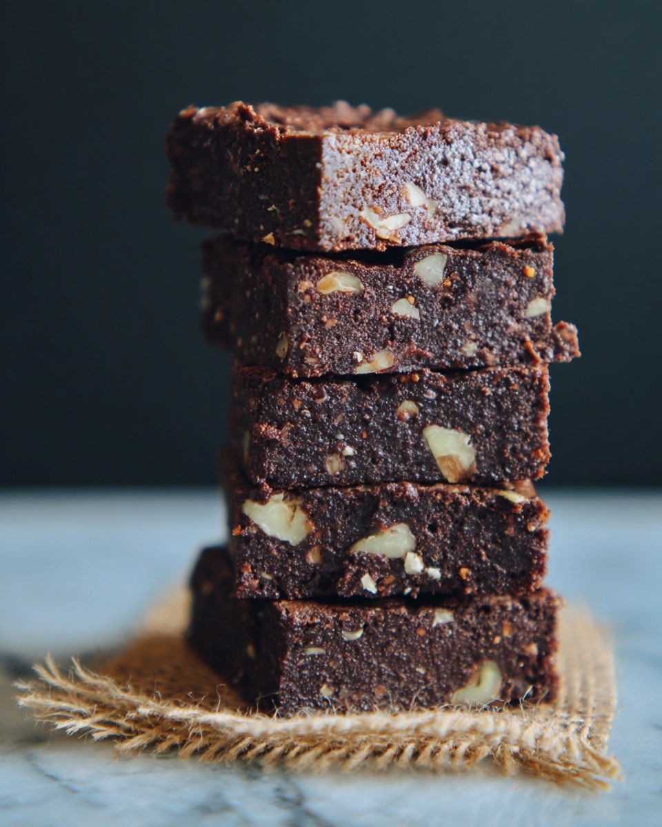 A tall stack of four square brownie pieces sits on a small piece of burlap on a white marbled surface. Each brownie layer is dark brown with a slightly rough texture, dotted with bits of light-colored nuts inside them. The brownies look thick and dense, and the edges are clean and straight. The background is simple and dark, making the stack stand out clearly. photo taken with an iphone --ar 4:5 --v 7