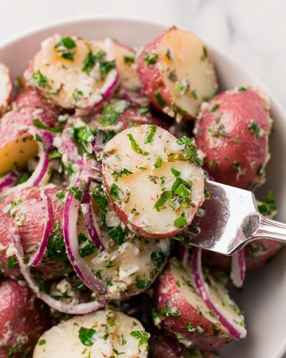 The image shows a close-up of a potato salad with several layers visible. The bottom layer is made of halved red-skinned potatoes with a smooth, creamy white inside and a light red-brown skin. Mixed throughout the potatoes are thin slices of purple-red onion, giving a pop of color and texture contrast. The entire salad is sprinkled with small pieces of fresh green herbs, possibly parsley, that add a bright green color across the surface. A silver fork lifts one potato half, showing its moist texture and the herbs sticking to it. The dish is placed on a white marbled surface. Photo taken with an iphone --ar 4:5 --v 7