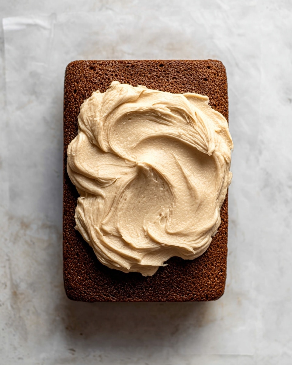 A single rectangular layer of dark brown cake with a smooth, slightly porous texture sits flat on a white marbled surface. On top at the center, there is one large dollop of light tan frosting, creamy and whipped with soft peaks and swirls showing movement, contrasting with the darker cake layer beneath it. Photo taken with an iphone --ar 4:5 --v 7