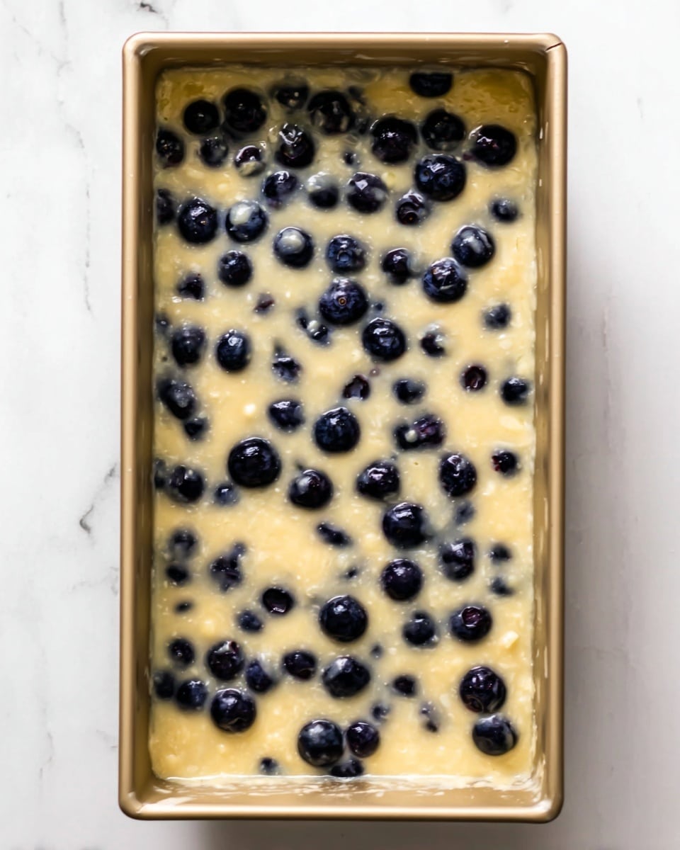 The image shows a rectangular baking pan filled with a batter that has many blueberries mixed in. The batter is light yellowish with a smooth and slightly thick texture. The blueberries are dark blue and scattered evenly throughout the batter, some partially sinking into it. The pan is on a white marbled surface. photo taken with an iphone --ar 4:5 --v 7