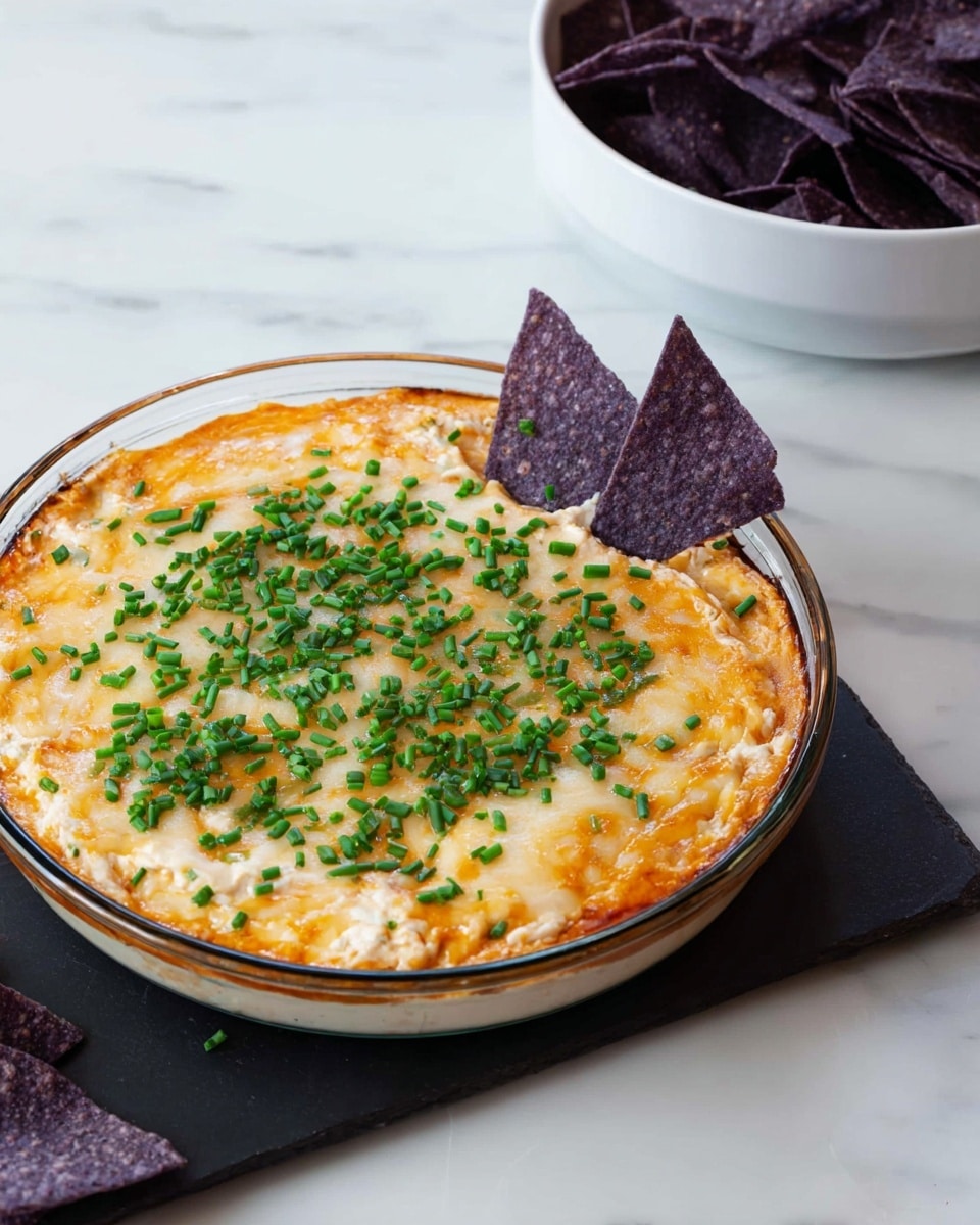 A round glass dish holds a layered dip with a melted light golden cheese top layer sprinkled with small chopped bright green chives, covering a creamy orange sauce underneath, and a pale creamy layer at the bottom, creating three distinct layers visible through the transparent dish. Two dark purple triangular chips are placed upright near the edge, partially inside the dish, and a white bowl filled with similar purple chips sits in the background on a white marbled surface. The scene looks clean and inviting with a simple setup photo taken with an iphone --ar 4:5 --v 7