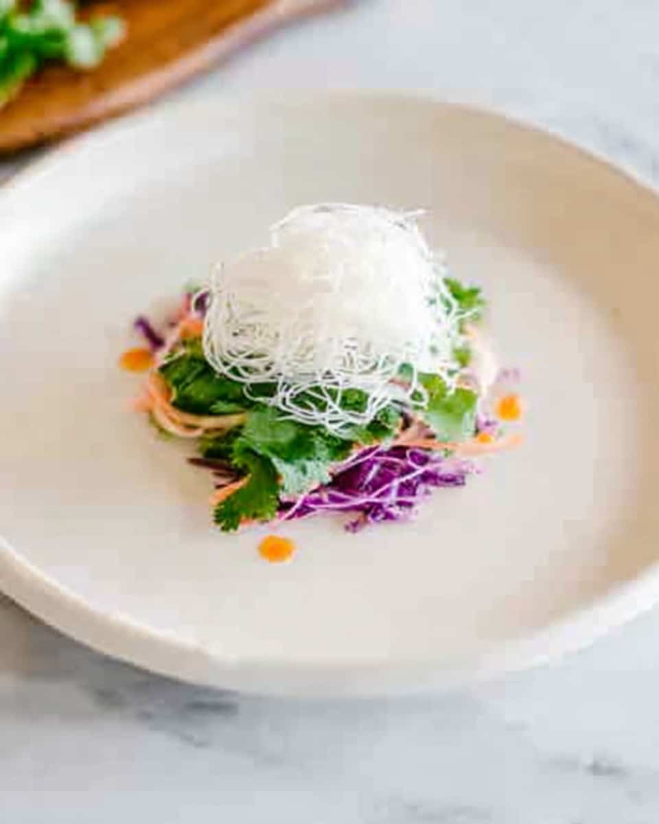 A white plate on a white marbled surface holds a small, neat stack of food in the center. The bottom layer looks like green cilantro leaves. On top of that, there is a small amount of thin, white noodles, shaped in a soft mound. Under the noodles, thin strips of bright purple cabbage and some orange sauce dots are visible, adding color to the dish. The food looks fresh and carefully arranged. photo taken with an iphone --ar 4:5 --v 7