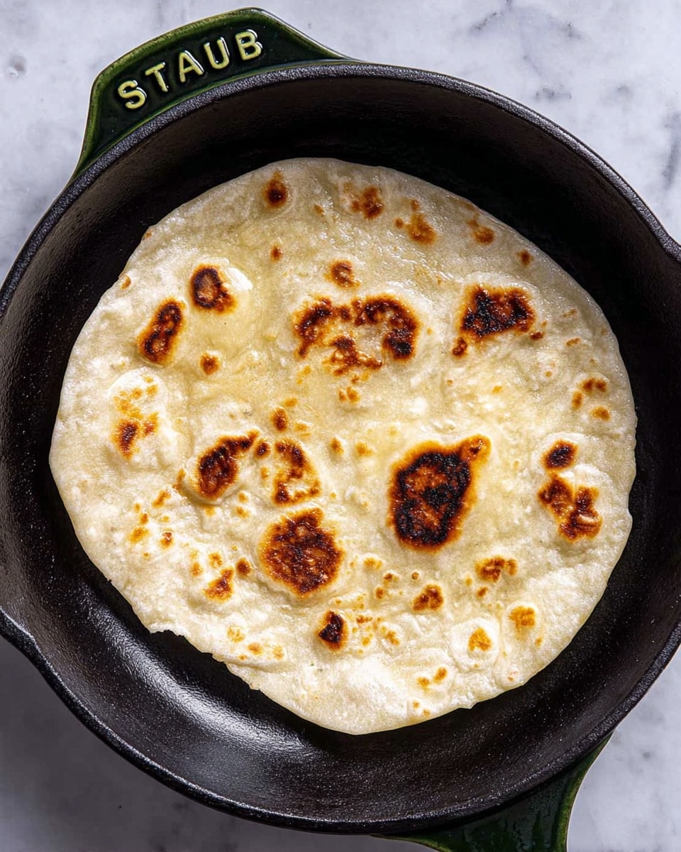 A single round flatbread with a soft, pale cream color and several dark brown toasted spots is cooking in a black cast iron pan. The flatbread has a slightly uneven edge and a bubbly texture on its surface. The pan has a dark green handle with the word