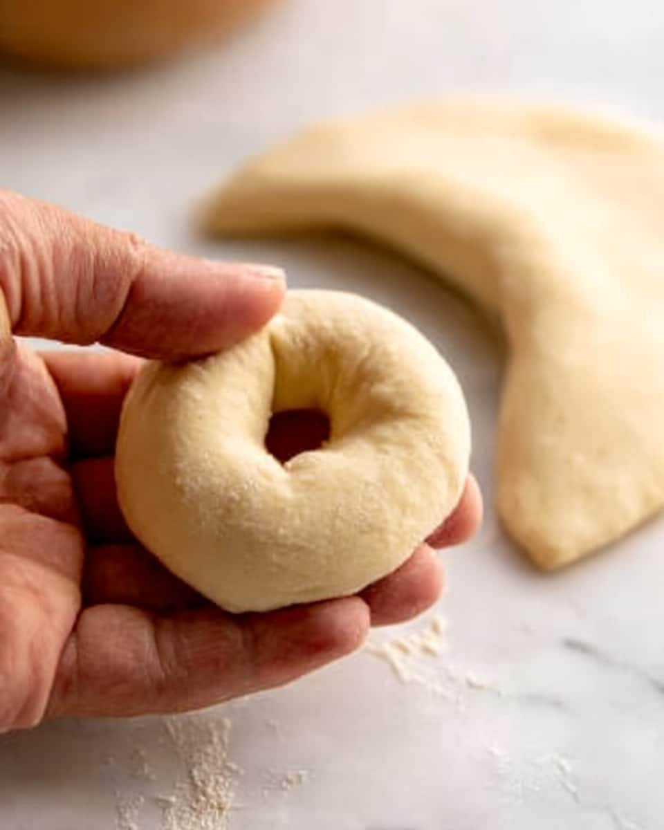 A woman’s hand is holding a small, smooth, round dough piece with a hole in the middle, like a small doughnut. The dough is light beige and soft. Behind it, on a white marbled surface, there is a bigger piece of rolled dough that is crescent-shaped, showing a flat, slightly textured surface. The photo is close up, focusing on the dough details. Photo taken with an iphone --ar 4:5 --v 7