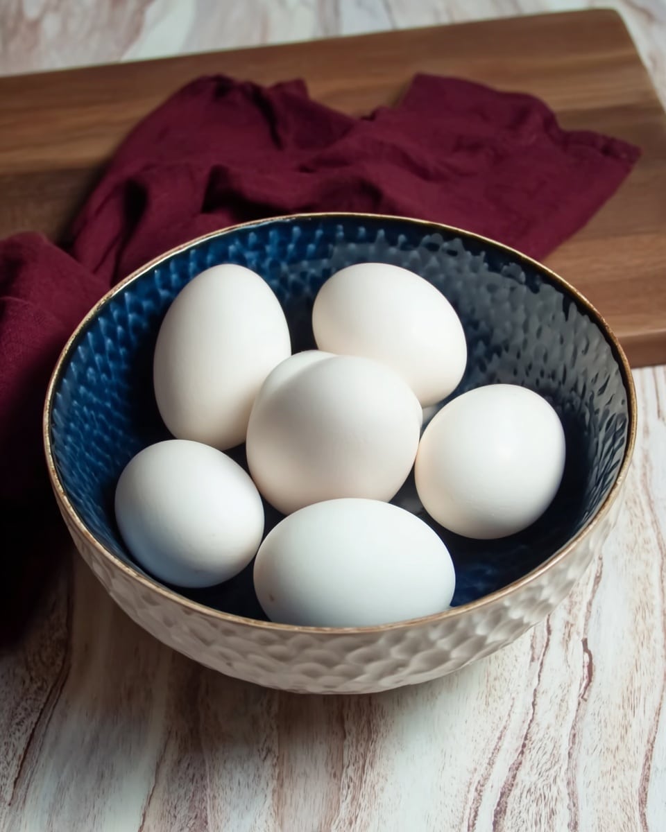 A white bowl filled with eight smooth white eggs resting inside, the bowl has a rippled texture on the outside and a deep blue inner surface with a gold rim. The bowl sits on a wooden surface with a white marbled texture, and behind it is a folded dark red cloth. The light softly highlights the eggs' shiny surfaces, showing their round shapes clearly photo taken with an iphone --ar 4:5 --v 7