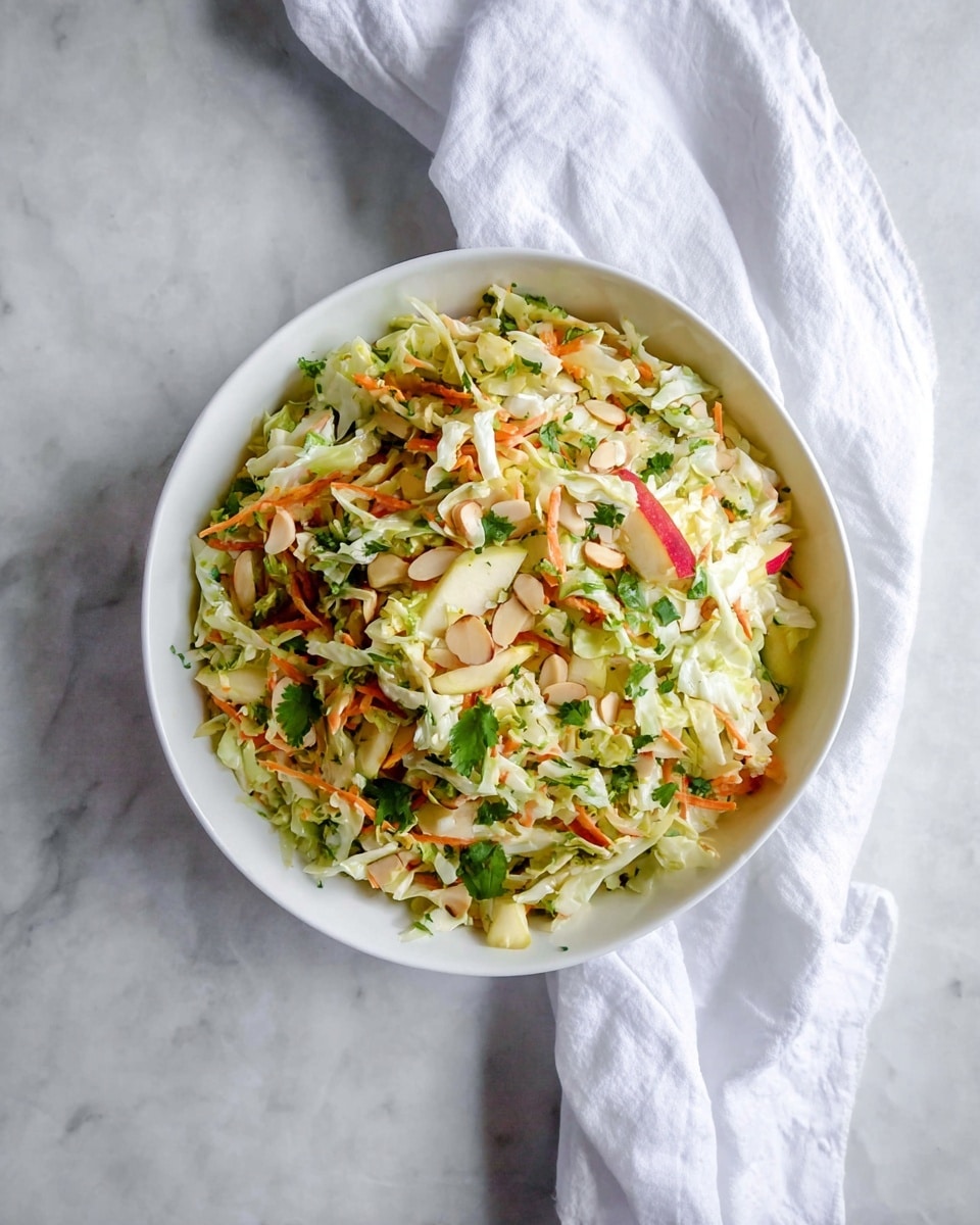 A white bowl filled with a colorful salad piled high, showing a mix of thin light green cabbage slices, small bright orange carrot shreds, and scattered pieces of light red apple with skin on. The salad is speckled with fresh green cilantro leaves and thin brown almond slices, giving texture and color contrast. The bowl sits on a white marbled surface with a white cloth casually placed on the side. The photo taken with an iphone --ar 4:5 --v 7