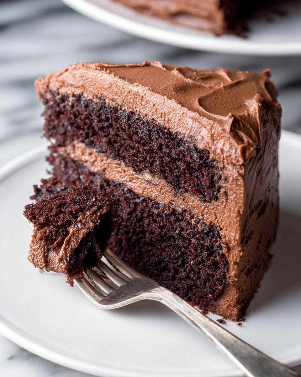 A close-up of a slice of chocolate cake on a white plate with a white marbled surface in the background. The cake has two thick layers of dark, moist chocolate sponge with a rich, smooth chocolate frosting layer in the middle. The outside of the cake is fully covered with the same creamy chocolate frosting. A silver fork holds a bite-sized piece of the cake at the front edge of the plate. The texture of the cake looks soft and dense, while the frosting appears light and fluffy. Photo taken with an iphone --ar 4:5 --v 7