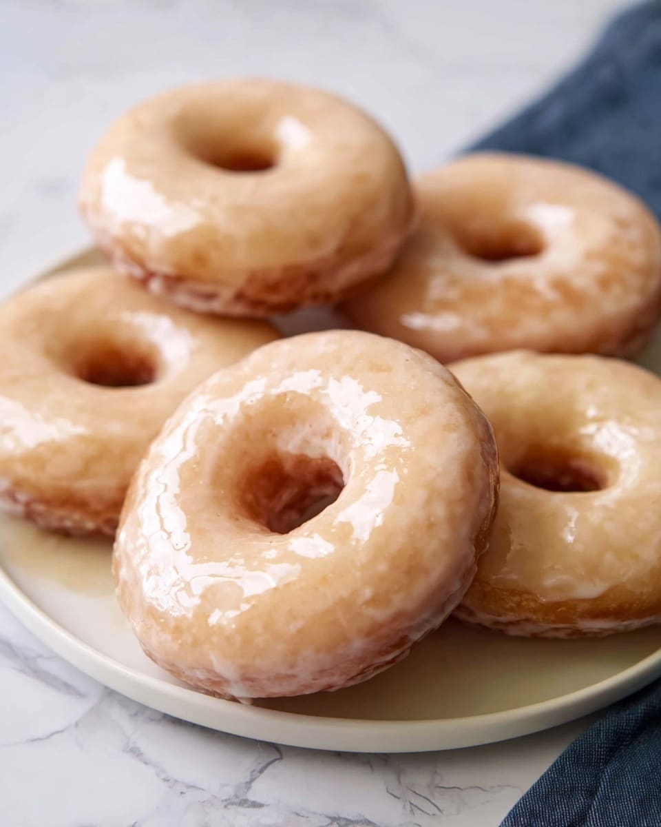 The image shows a white plate with five glazed donuts placed on it. Each donut has a shiny, smooth, light golden glaze covering the entire surface, creating a slightly wet look. The donuts have a soft texture with a classic round shape and a hole in the middle. The plate sits on a white marbled surface, adding a clean and bright background to the simple arrangement of donuts. photo taken with an iphone --ar 4:5 --v 7