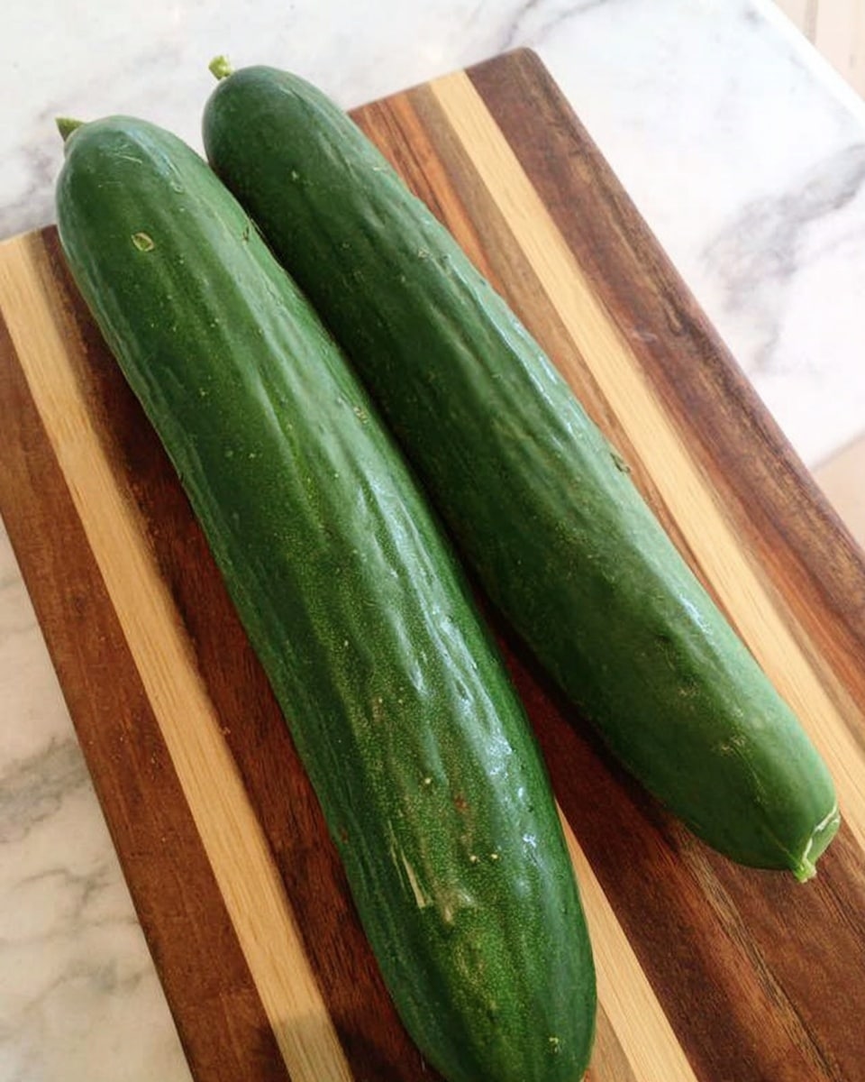 Two long, dark green cucumbers with a slightly shiny and textured skin lie side by side on a wooden cutting board with light and medium brown stripes, placed on a white marbled surface. The cucumbers are fresh, smooth, and have a firm look, with one slightly thicker than the other. No other items or objects are seen around. Photo taken with an iphone --ar 4:5 --v 7
