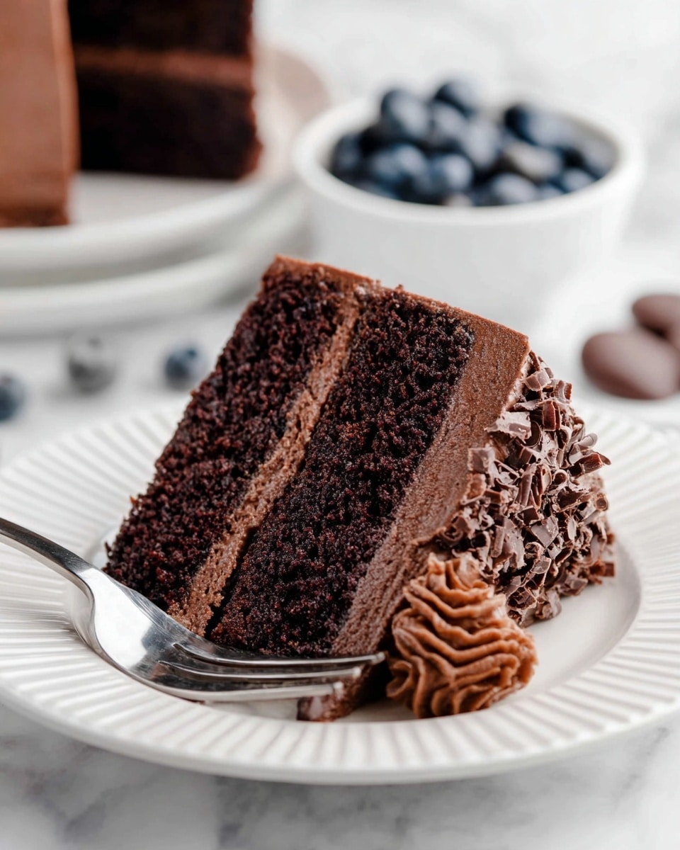 This image shows a round chocolate cake with smooth, rich dark brown frosting covering all sides. On top, there is a dark chocolate swirl in the middle, surrounded by eight evenly spaced dollops of darker piped chocolate cream decorated with chocolate shavings. The cake is placed on a white marbled surface. In the background, there is a white bowl with chocolate chunks on the left, pink flowers behind the cake, and a white pedestal bowl filled with bright red raspberries on the right. Photo taken with an iphone --ar 4:5 --v 7