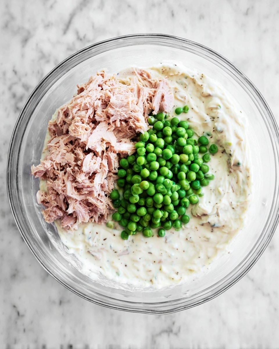 A clear glass bowl holds three main layers of food ready to be mixed. The bottom layer is a thick, creamy white sauce with small black specks, covering most of the bowl's inside. On top, near the upper part of the bowl, there is a light pink, shredded meat layer that looks soft and moist. Below the meat and slightly overlapping it, a pile of small, round bright green peas sits in the center of the bowl. The bowl is placed on a white marbled surface. photo taken with an iphone --ar 4:5 --v 7