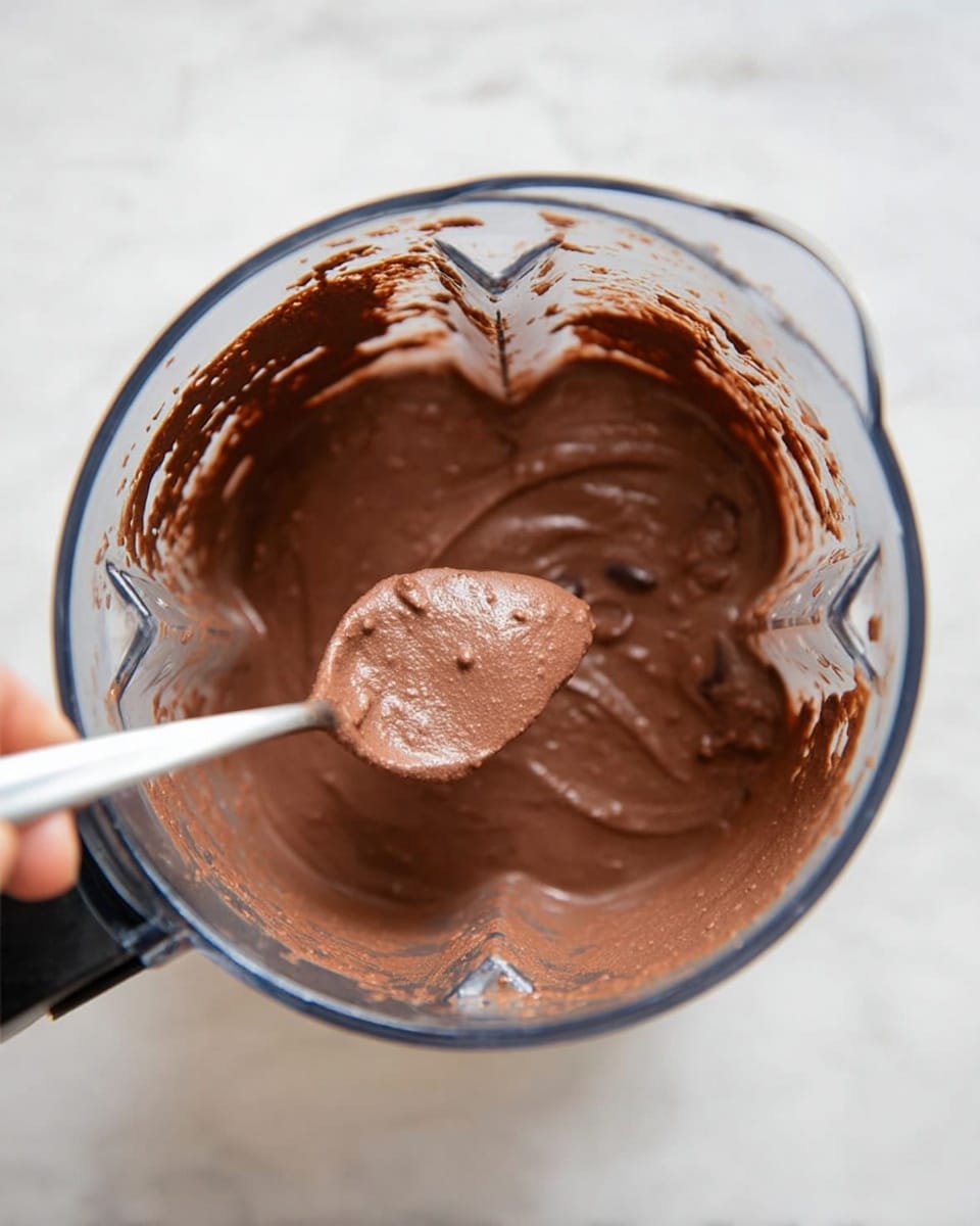The image shows a clear blender jar filled with smooth, creamy chocolate batter. The batter is rich brown and thick, clinging to the sides of the jar with some splatters at the top edges. A spoon covered in the chocolate batter is held above the jar by a woman's hand, showing the texture closely. The background is a white marbled surface. photo taken with an iphone --ar 4:5 --v 7
