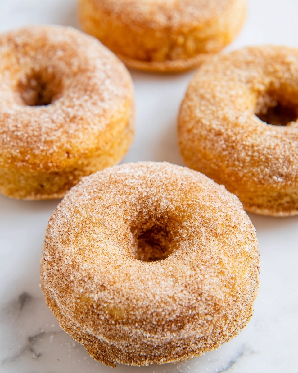 The image shows five round donuts with a hole in the center, arranged on a white marbled surface. Each donut is golden brown with a slight rough texture, covered evenly with a light dusting of fine white sugar and a touch of cinnamon spice, giving a soft powdery look on the surface. There are no other colors or toppings; the focus is on the simple, sweet coating contrasting with the warm golden color of the dough. The donuts sit close but not touching, with the main one in the center and four around it. Photo taken with an iphone --ar 4:5 --v 7