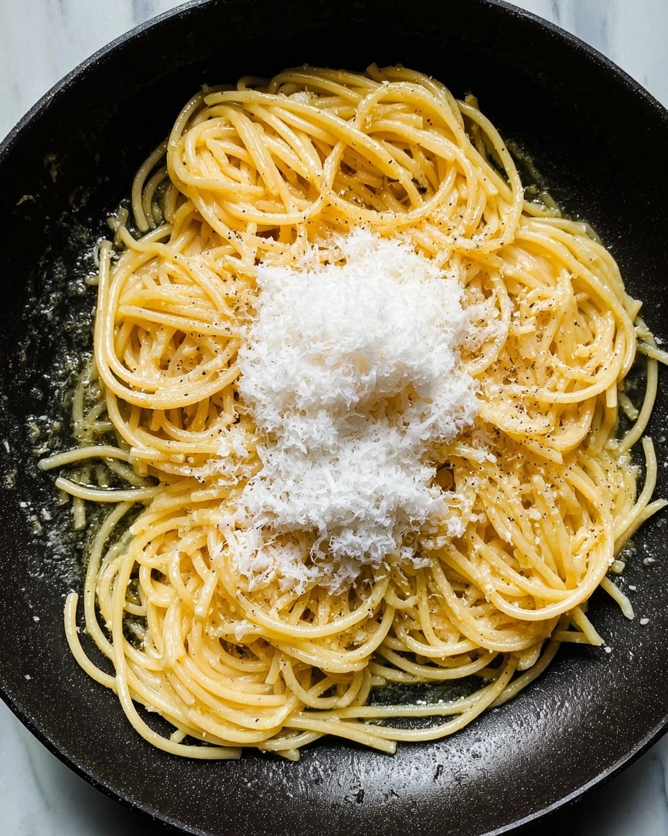 A close-up view of a black pan filled with round spaghetti noodles arranged in a loose heap, coated lightly with olive oil and specks of cracked black pepper. In the center, there is a tall mound of finely grated white cheese, slightly melting over the warm pasta. The surface under the pan is a white marbled texture. photo taken with an iphone --ar 4:5 --v 7