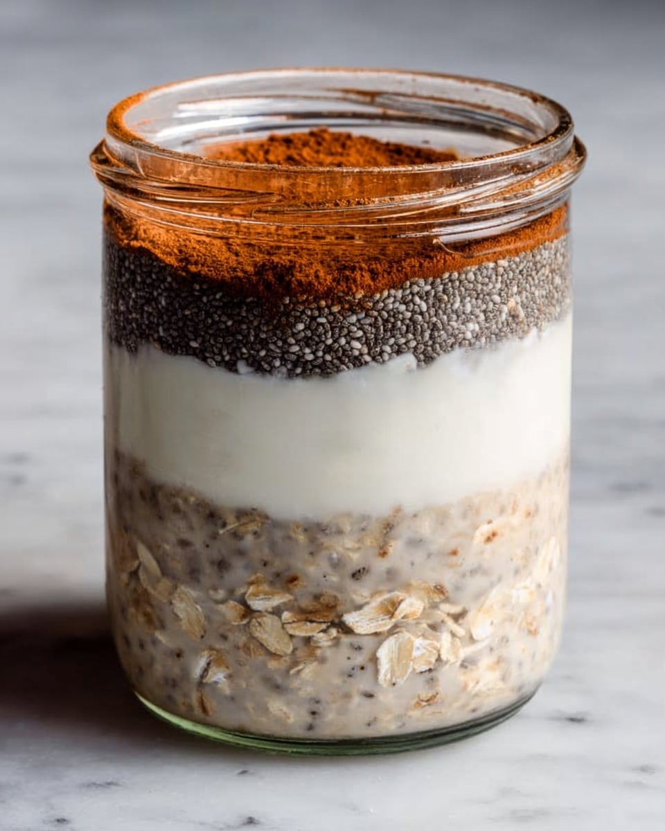 A clear glass jar is filled with a layered mix of soaked oats at the bottom, creamy white yogurt or milk with visible oat pieces in the middle, topped with a dark layer of chia seeds, and finished with a dusting of light brown cinnamon powder on the very top. The jar is placed on a white marbled surface, showing the textures and colors through the glass clearly. photo taken with an iphone --ar 4:5 --v 7