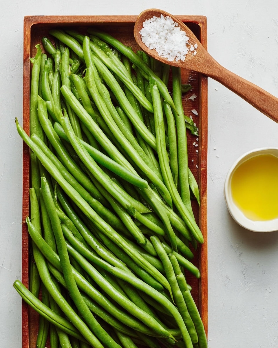 A rectangular wooden tray filled with fresh green beans laid flat in neat layers, showing their smooth, vibrant green texture and long, thin shapes. To the right of the tray is a small white bowl with a yellow liquid inside, partially visible, and above it is a wooden spoon with coarse white salt crystals resting on a white marbled surface. The background is clean and bright, enhancing the fresh look of the green beans. photo taken with an iphone --ar 4:5 --v 7