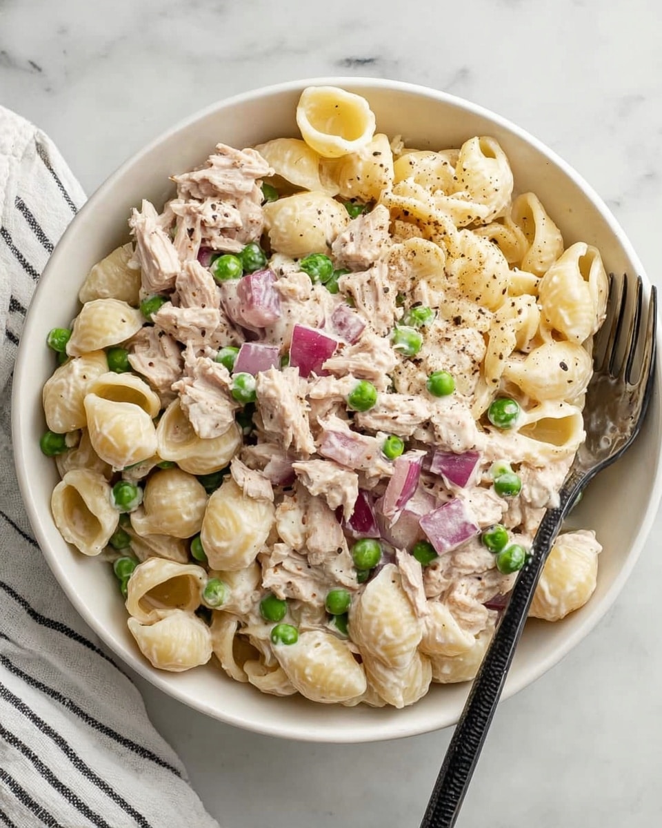 A white bowl filled with a creamy pasta salad made of shell pasta, light beige in color, mixed with small green peas and chopped purple onions. The pasta is fully coated in a smooth white sauce that looks thick but soft, with some specks of black pepper on top. The salad has visible chunks of light beige tuna scattered evenly throughout. A shiny silver fork rests inside the bowl on the right side, and the bowl is placed on a white marbled surface with a black and white striped cloth to the left. photo taken with an iphone --ar 4:5 --v 7