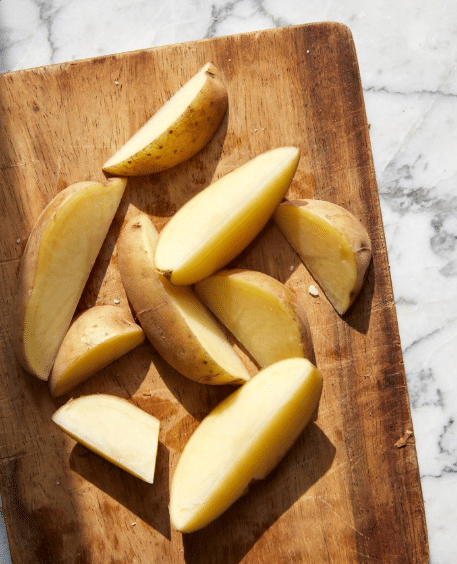 Several potato wedges are placed on a wooden cutting board with a white marbled surface nearby. The wedges show a light brown skin on one side and smooth, pale yellow flesh on the other. The wedges vary in size but mostly have a thick and firm texture with pointed ends. Photo taken with an iphone --ar 4:5 --v 7