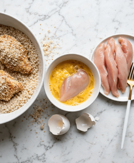 The image shows three white bowls arranged on a white marbled surface. On the left, a large bowl filled with light beige breadcrumbs mixed with sesame seeds contains two pieces of chicken coated with the crumbs, showing a rough textured surface. On the right, a white plate holds several raw chicken strips with a pale pink color and glossy texture, placed next to a fork with a white handle and a gold neck. In the center, a smaller bowl contains a yellow beaten egg mixture, partially covering a single piece of raw chicken. Two pieces of broken white eggshells lie nearby on the white marbled background. Photo taken with an iphone --ar 4:5 --v 7