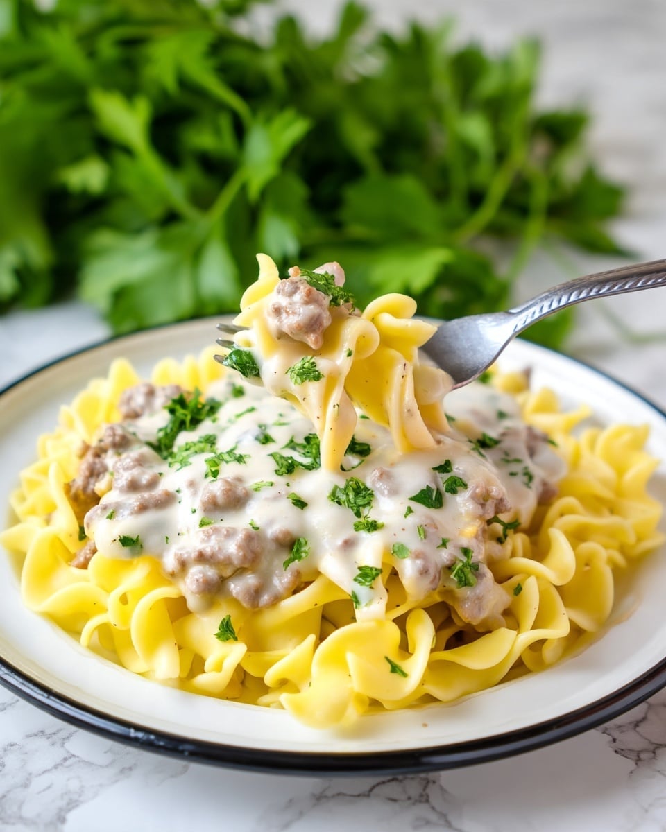 A white plate holds a dish with three layers; the bottom layer is light yellow egg noodles with soft, wavy edges. The middle layer is a thick white cream sauce mixed with small browned meat pieces and bits of onions spread evenly on top of the noodles. The top layer has small bits of finely chopped green herbs sprinkled softly over the sauce. A woman's hand with pale skin and neatly trimmed nails holds a shiny silver fork, digging into the noodles and sauce from the top right. In the background, there is a blurred bunch of green parsley and a brown pumpkin, all set on a white marbled surface. Photo taken with an iphone --ar 4:5 --v 7