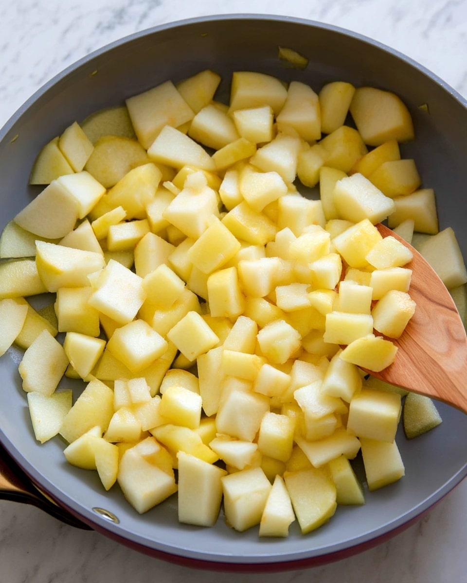 The image shows a grey pan filled with many small pieces of peeled apples, all light yellow and soft in texture, spread out evenly inside. A wooden spoon is partially inside the pan on the right side, slightly lifting some apple pieces, showing a mix of small square and triangular shapes. The background surface is white marbled texture. photo taken with an iphone --ar 4:5 --v 7