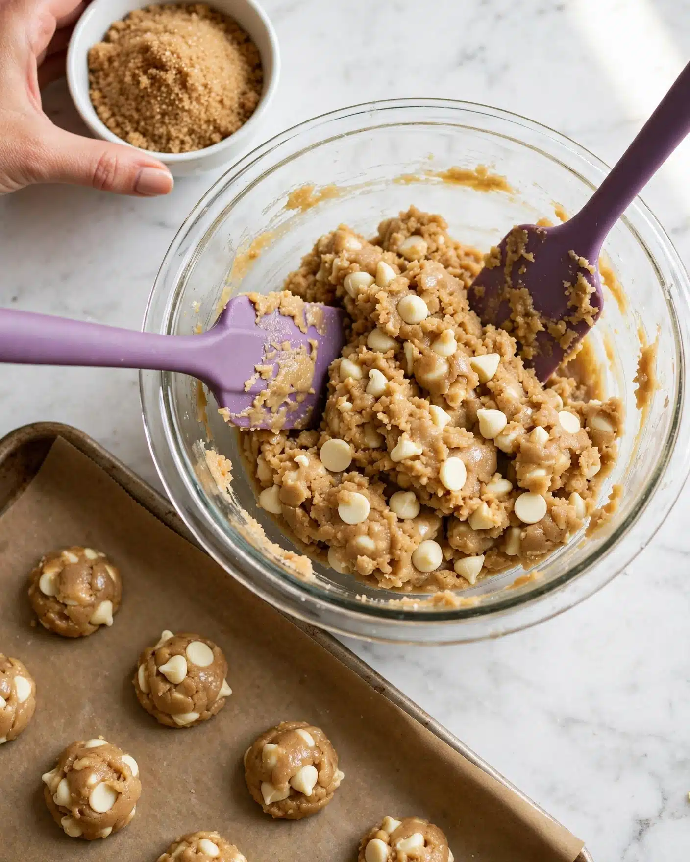 A clear glass bowl filled with thick light brown cookie dough mixed with white chocolate chips sits on a white marbled surface. Two purple spatulas rest inside the bowl, one on the left side and one on the right side, partially covered with dough. In the top left corner, a woman's hand holds a small white bowl with brown sugar, poised above a baking sheet lined with brown parchment paper. On the parchment paper, there are evenly spaced small dollops of dough, ready to be baked. Photo taken with an iphone --ar 4:5 --v 7