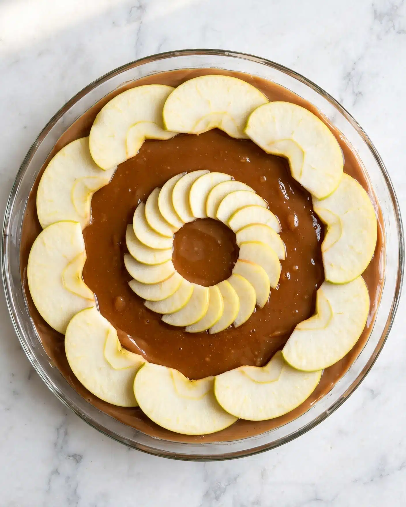 Caramel Apple Upside Down Cake Recipe 4 A round clear glass dish holds a layered dessert starting with a smooth, light brown syrup layer at the bottom. On top, there are thin apple slices arranged in two circular rings; the outer ring has large pale yellow apple slices evenly spaced and slightly overlapping each other, while the inner ring contains smaller apple slices similarly fanned out. The dish is placed on a white marbled surface. The overall look is neat and orderly with soft colors and clear details photo taken with an iphone --ar 4:5 --v 7