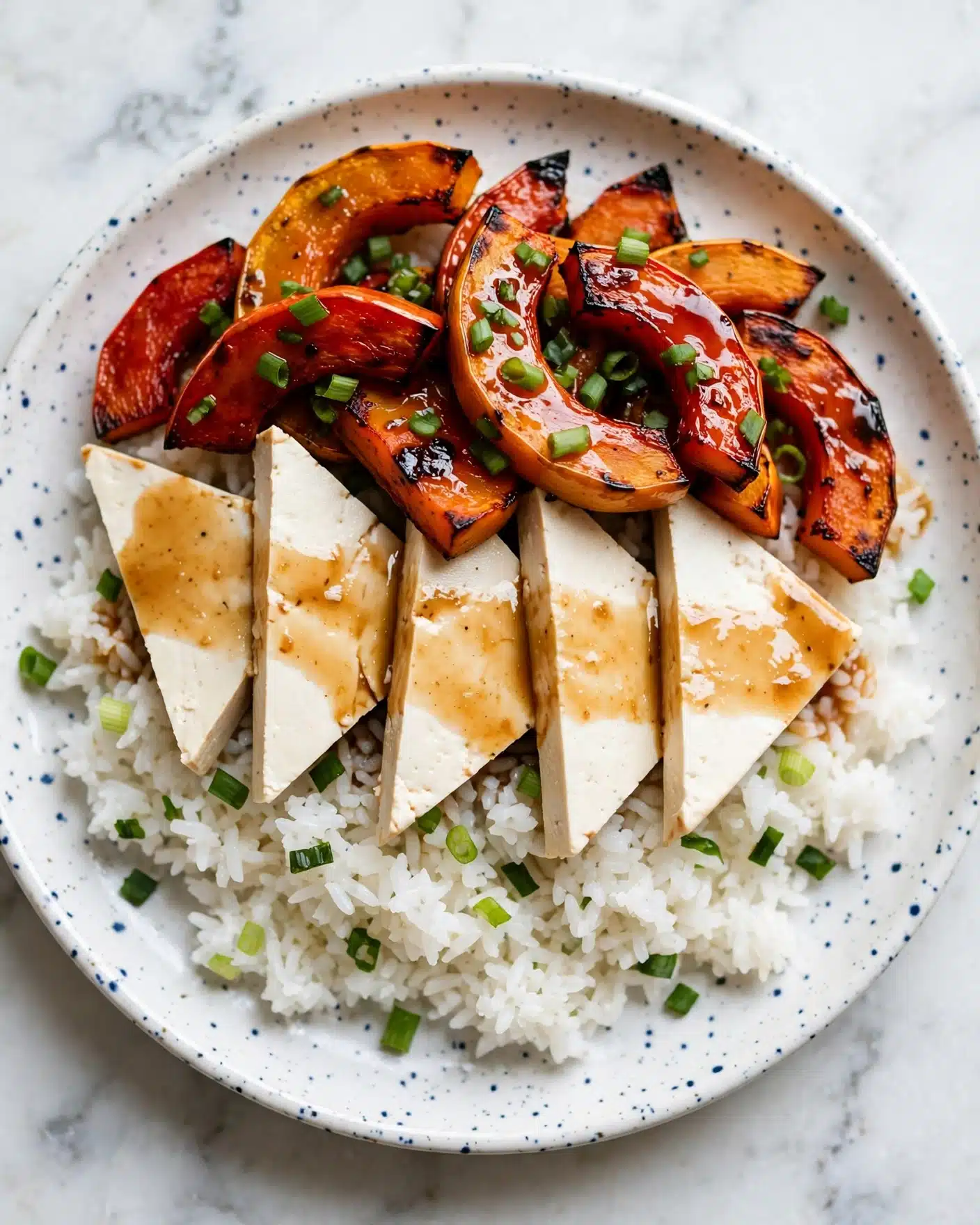 The dish shows a white round plate placed on a round wooden board, filled with a stack of roasted acorn squash rings. There are about two layers of these orange to deep brown colored squash rings, with a caramelized look and a shiny texture from a glaze or sauce. The squash rings are arranged unevenly, some overlapping each other. Scattered small white sesame seeds add a touch of contrast, and fresh green herb leaves are sprinkled across the top and around the plate for color. The background is a white marbled surface, and a white cloth is partly visible above the plate. photo taken with an iphone --ar 4:5 --v 7