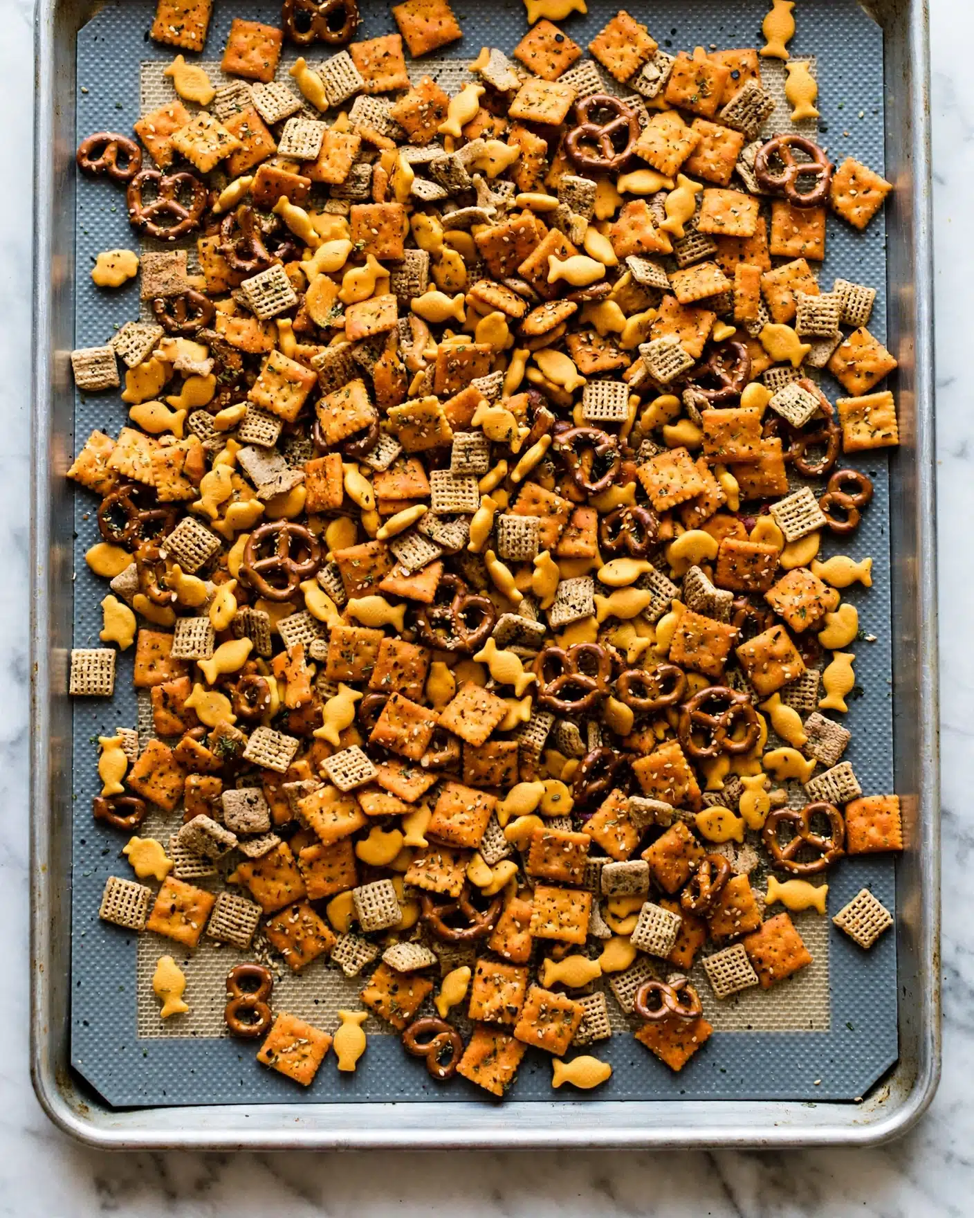 A close-up view of a snack mix spread over a baking tray with a textured mat beneath. The mix contains several types of small snacks: square orange crackers, small yellow fish-shaped crackers, dark brown pretzels, and small light brown square cereal pieces. The entire mix is covered with a seasoning that looks like sesame seeds and herbs, adding a speckled texture across all the snacks. The color palette is mainly orange, brown, and yellow, with the seasoning creating small dark and light spots, and the snack pieces lie in an uneven layer. The tray is on a white marbled surface. photo taken with an iphone --ar 4:5 --v 7