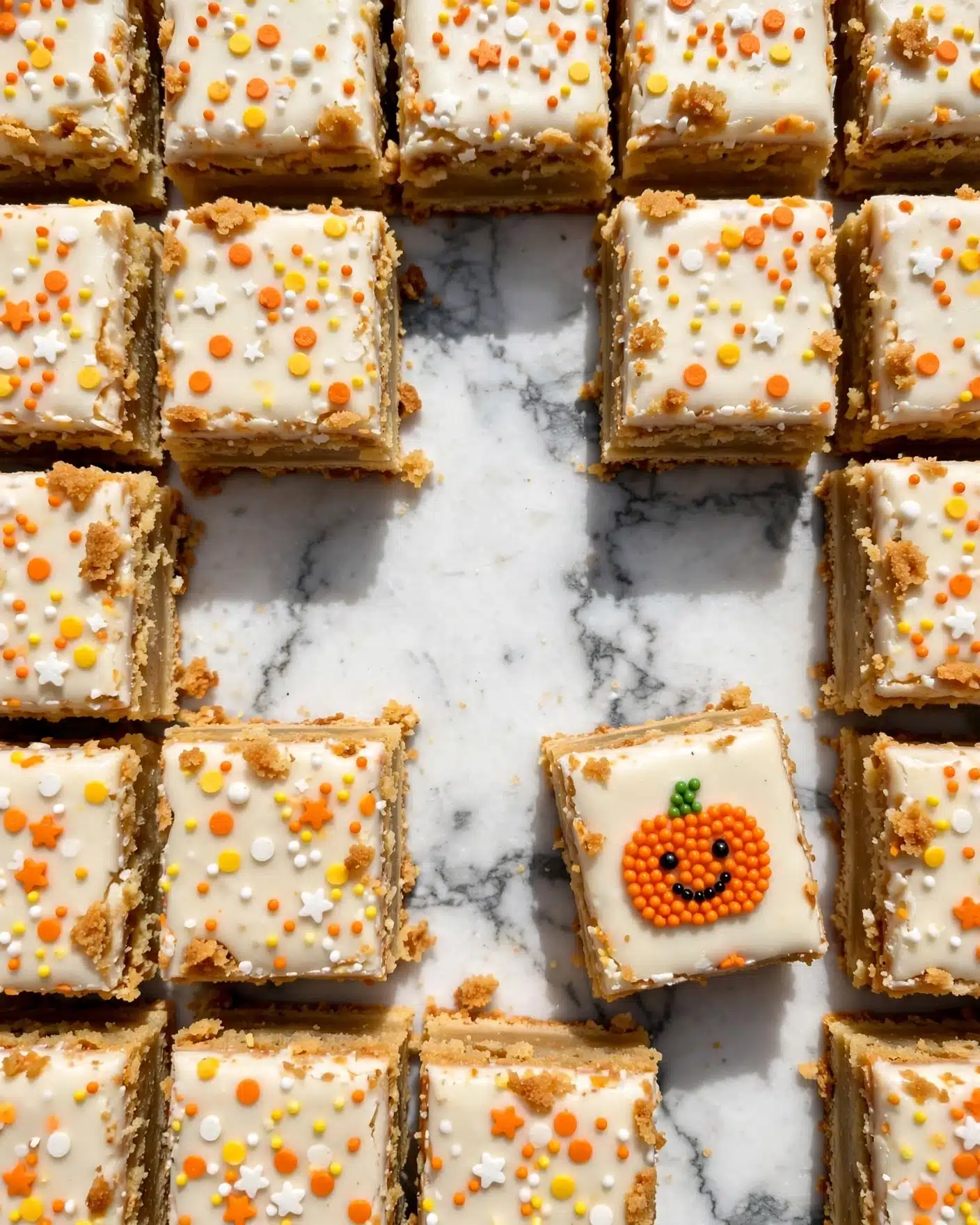 The image shows a close-up of square cake bars arranged in a grid pattern on a white marbled surface, with some pieces missing in the middle, creating gaps. Each cake bar has two layers: a thick, golden brown base layer with a slightly crumbly texture, topped with a smooth, creamy white icing layer. The icing is decorated with small colorful sprinkles in orange, yellow, and white, including stars and dots. One cake square stands out with a small orange pumpkin decoration made of tiny beads on the icing, smiling with black eyes and a green stem. The photo is bright and clear, showing texture in both the cake and icing. Photo taken with an iphone --ar 4:5 --v 7