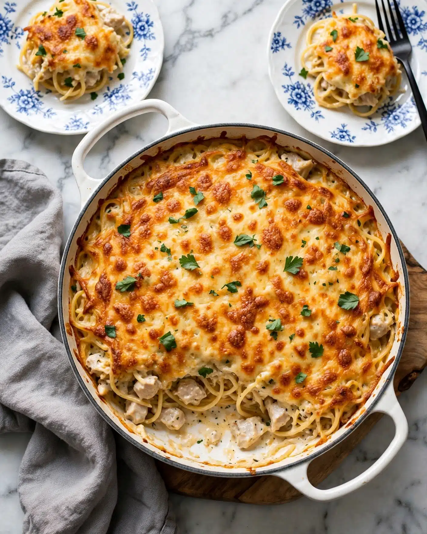 A white cast iron pan filled with baked spaghetti casserole that has a golden brown, melted cheese top layer with scattered small green herb leaves on it. The casserole's middle layer shows creamy spaghetti noodles mixed with chunks of white meat, lightly coated in sauce. Around the pan, you can see a wooden board underneath and a soft textured gray towel on one side. The background surface is white with a marbled texture. In the top corners, there are two white plates with blue floral designs holding small servings of the same spaghetti dish and a black fork resting on one plate. Photo taken with an iphone --ar 4:5 --v 7