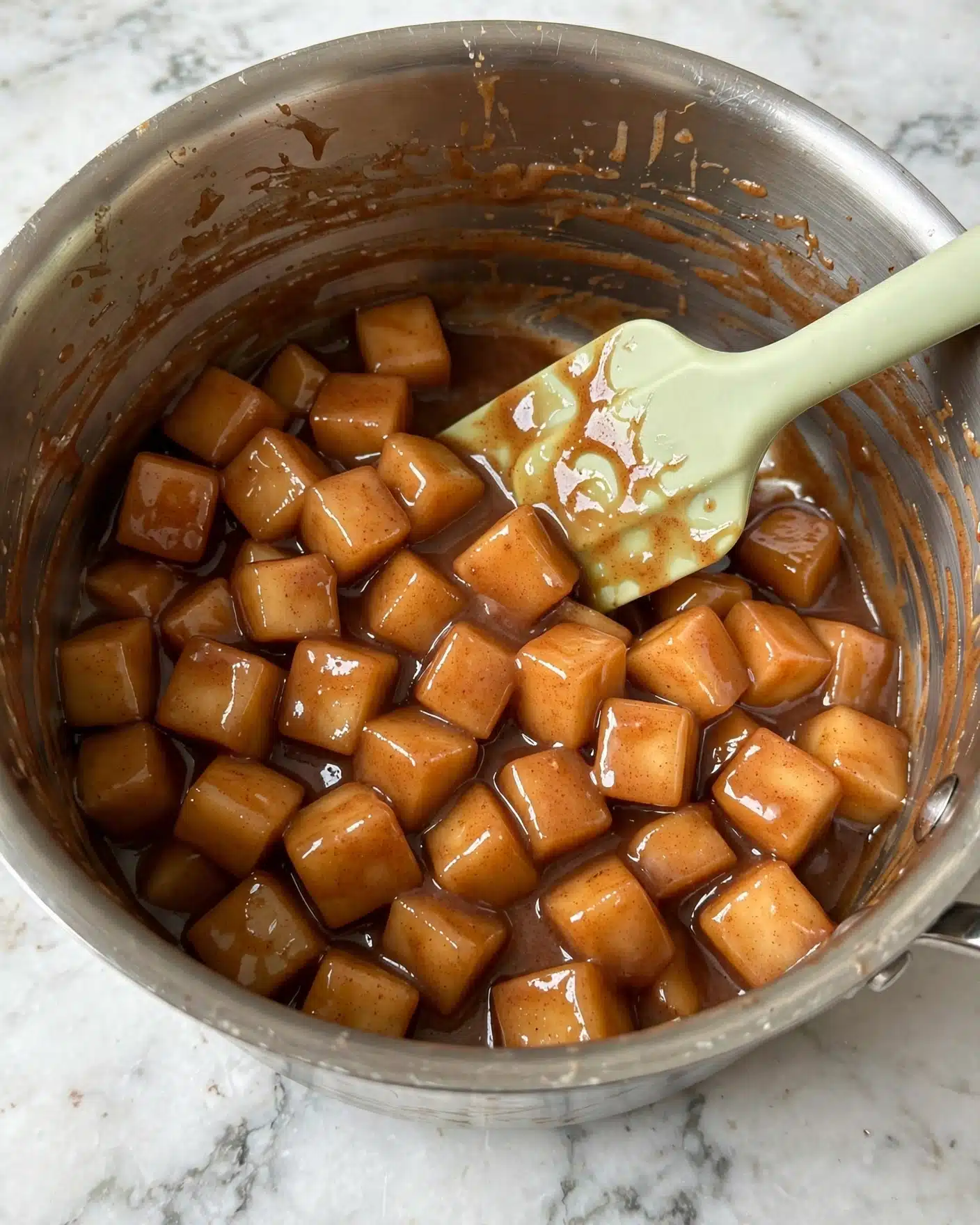Inside a silver pot on a white marbled surface, there are many small cubes of soft cooked apples coated in a glossy cinnamon-brown sauce. The apple pieces are light brown with a warm spice color blending evenly on each cube. A pale green spatula with a smooth texture rests inside the pot, partially covered in the thick, shiny sauce, mixing the apple cubes. The sides of the pot are slightly stained with the cinnamon sauce, showing recent stirring. Photo taken with an iphone --ar 4:5 --v 7