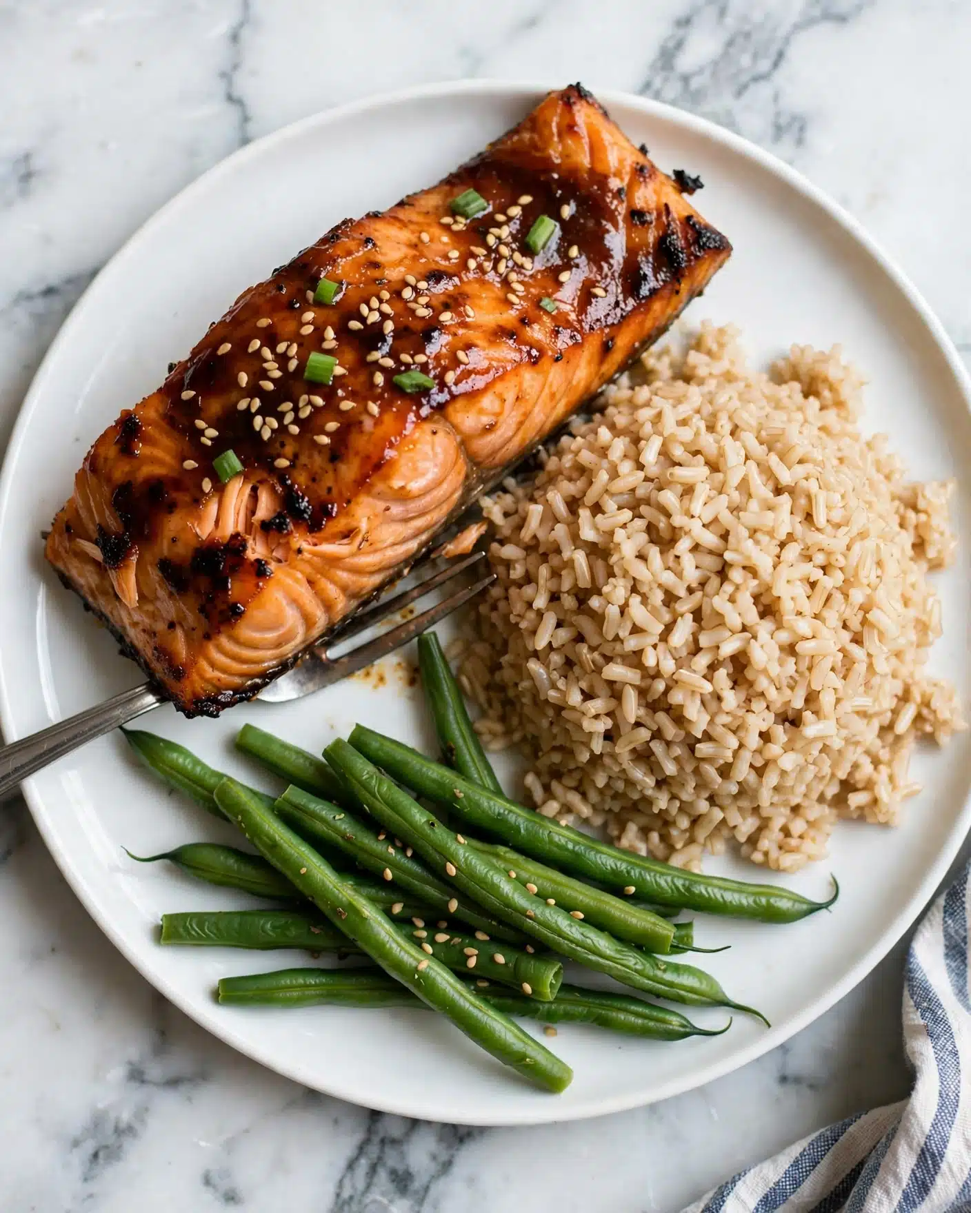 The dish shows a white plate with three main parts: a large piece of cooked salmon covered in a shiny, dark brown sauce sprinkled with light tan sesame seeds and small bits of green onions on top. The salmon has a textured, flaky orange surface with some darker charred edges, and a silver fork is partially under the salmon. To the right of the salmon is a generous mound of light brown cooked rice with a slightly uneven texture. Below the rice are green beans with a bright green color and a few sesame seeds scattered on them. The plate is set on a white marbled surface with part of a striped cloth visible near the bottom right corner. Photo taken with an iphone --ar 4:5 --v 7