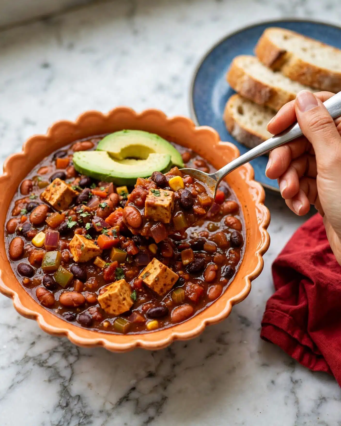Vegetarian Pumpkin Chili Recipe 5 A bowl of thick chili with a rich red-brown sauce filled with visible pieces of beans, chopped vegetables, and chunks of tofu or similar protein. The chili is presented in a scalloped-edged orange bowl, topped with two slices of green avocado. A woman's hand holds a spoon inside the bowl, scooping some chili. The bowl sits on a white marbled surface beside a blue plate with bread slices and a red cloth napkin. The colors and textures of the chili look hearty and freshly cooked. Photo taken with an iphone --ar 4:5 --v 7