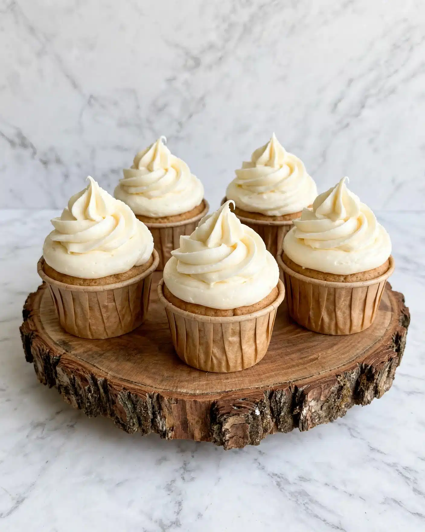 The image shows five cupcakes on a wooden stand with rough bark edges. Each cupcake has one base layer that is light brown and textured, wrapped in a light brown paper liner. On top of each cupcake is one thick layer of smooth, creamy off-white frosting swirled in a high peak shape. The background is a white marbled texture that contrasts softly with the cupcakes and wooden stand. photo taken with an iphone --ar 4:5 --v 7