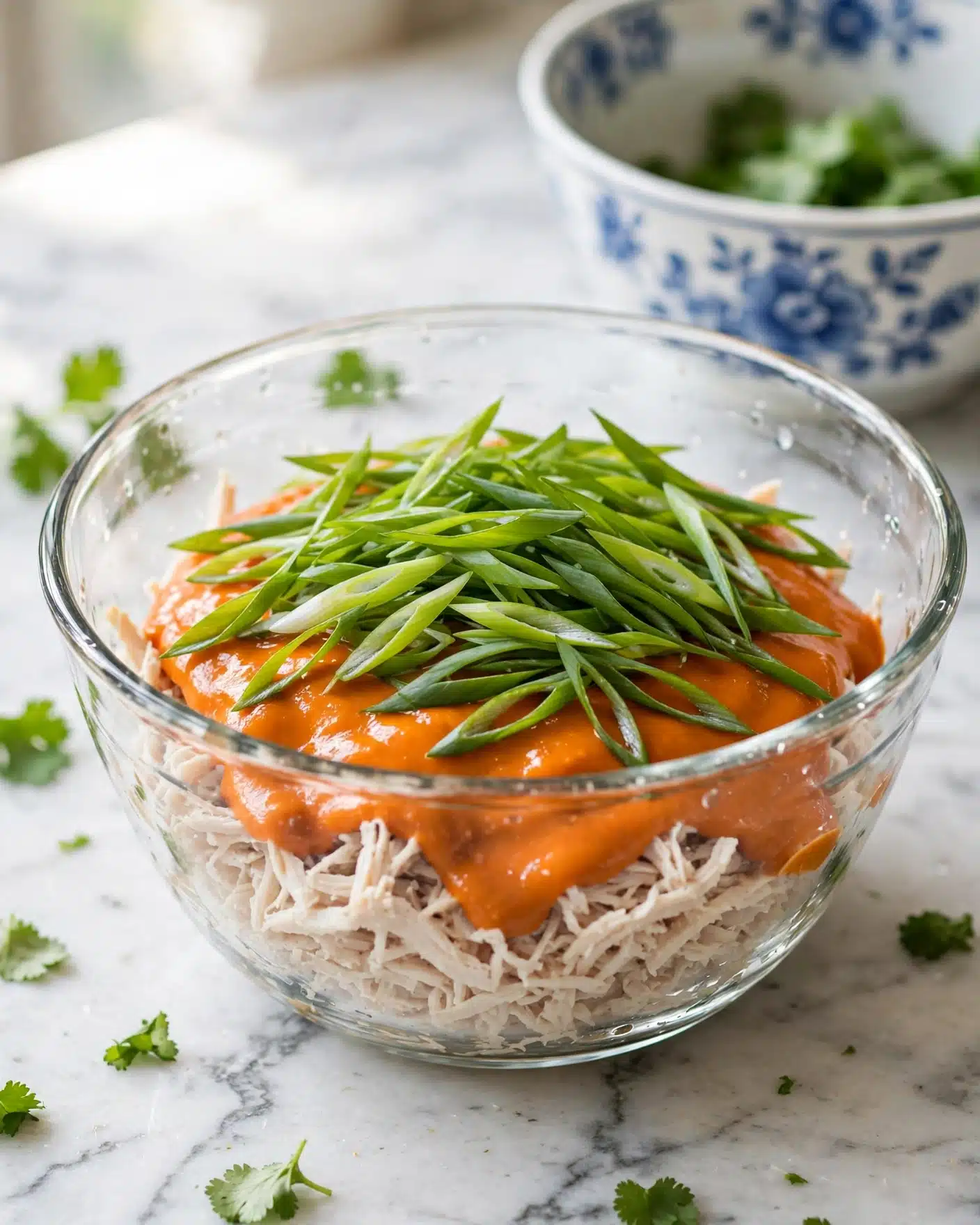 The image shows a clear glass mixing bowl with shredded white meat at the bottom, topped with a thick orange sauce. On top of the sauce, there is a generous layer of sliced green onions that are long and thin, spread evenly around the bowl. The bowl is on a white marbled surface with some scattered green leaves nearby, and there is a white bowl with blue flower patterns in the background partially filled with similar green leaves. Light shines softly onto the bowl, highlighting the fresh ingredients and creating clear reflections on the glass. Photo taken with an iphone --ar 4:5 --v 7