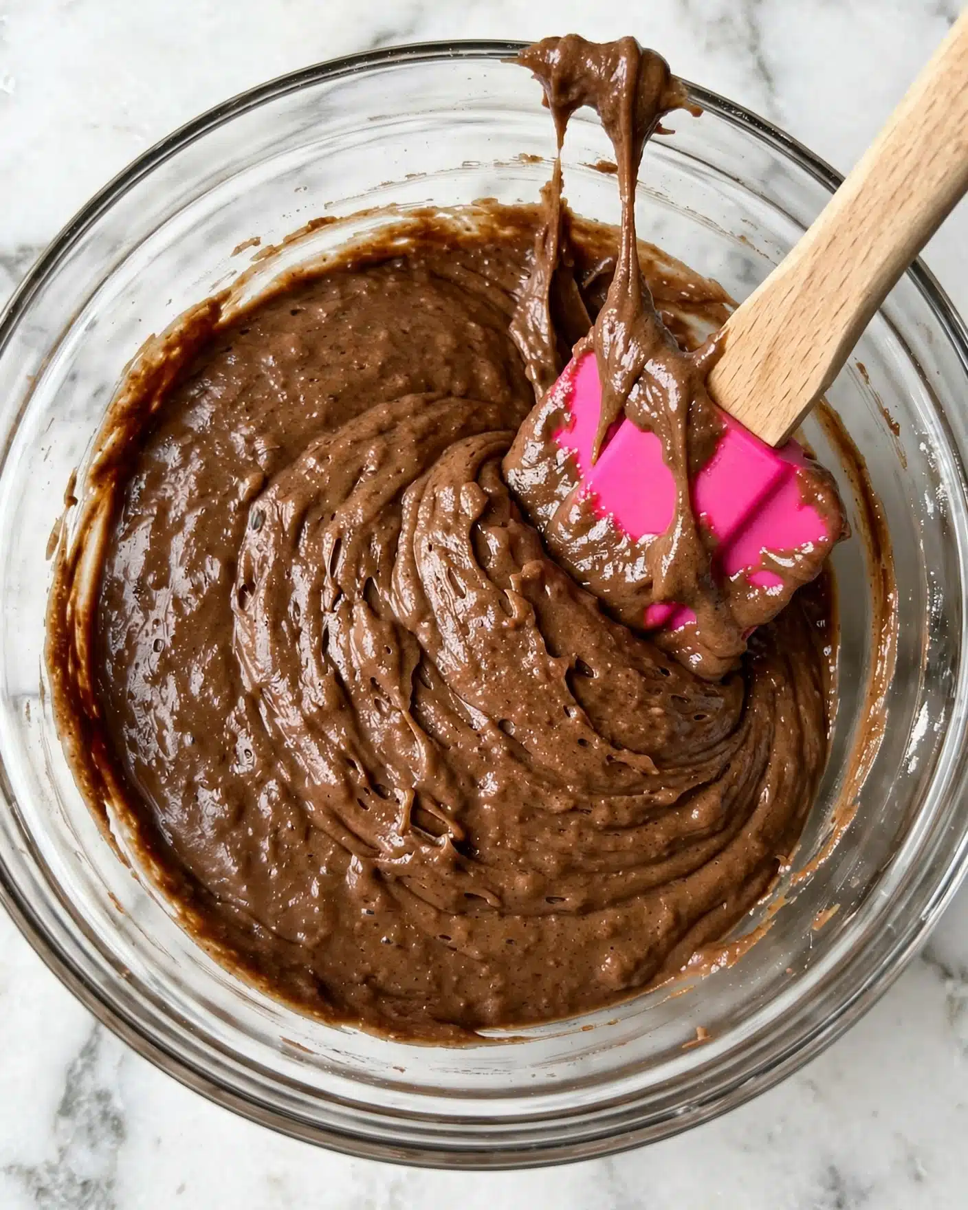 A close-up view of a single-layer thick brown batter with a smooth and slightly textured surface inside a clear glass bowl. A wooden spatula with a bright pink silicone tip is partially dipped into the batter, lifting some up with visible batter clinging to the tip. The bowl sits on a white marbled surface, providing a clean and simple background. Photo taken with an iphone --ar 4:5 --v 7