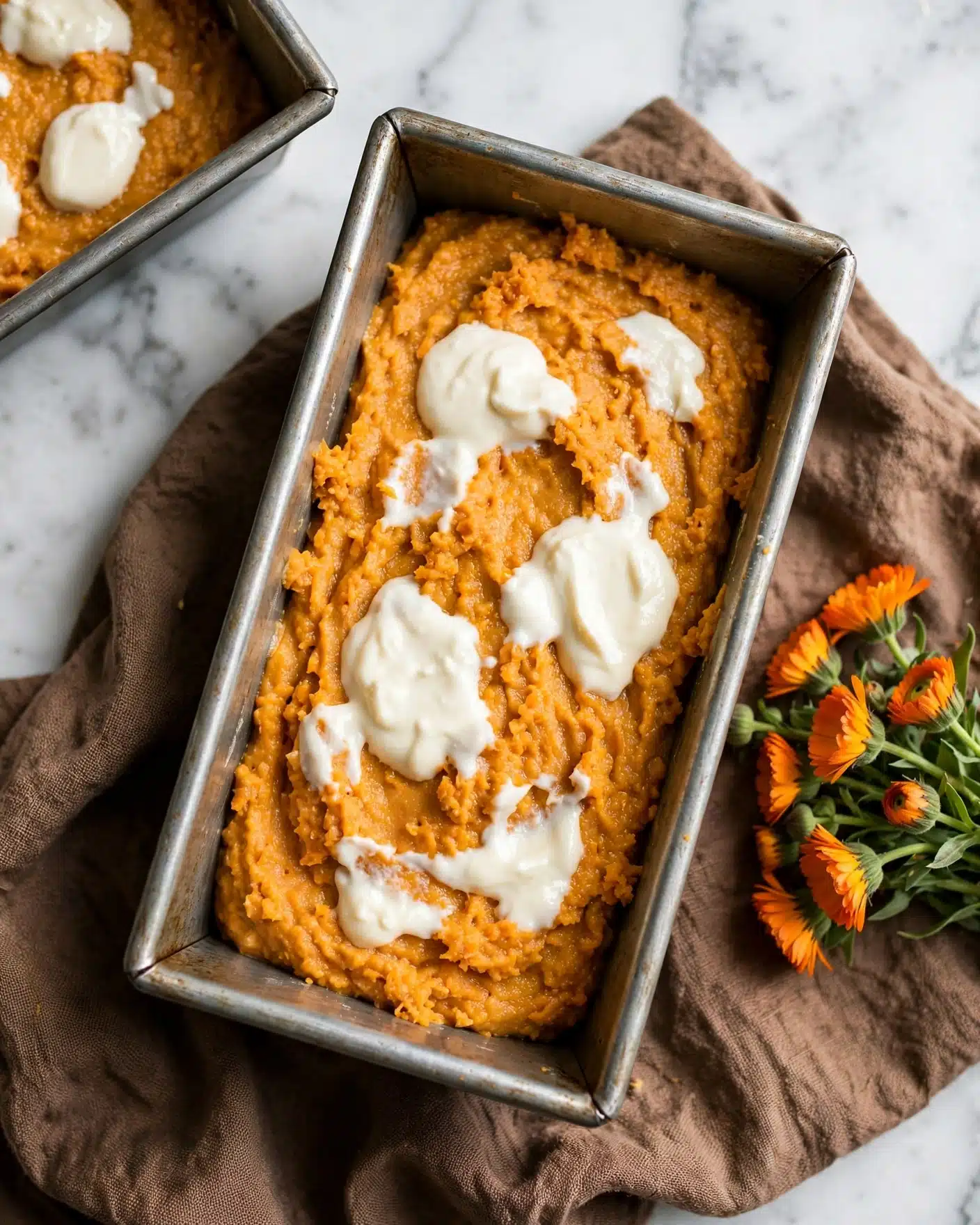Cream Cheese Swirled Cinnamon Pumpkin Butter Bread Recipe 4 The image shows a single metal loaf pan filled with a thick, orange batter that looks like pumpkin or carrot cake batter. On top of this batter, there are dollops of white cream spread unevenly across the surface. The pan is placed on a brown textured fabric, with a small bunch of orange flowers nearby. In the upper left corner, part of another metal loaf pan with similar batter and cream is visible. The background is a white marbled texture. photo taken with an iphone --ar 4:5 --v 7