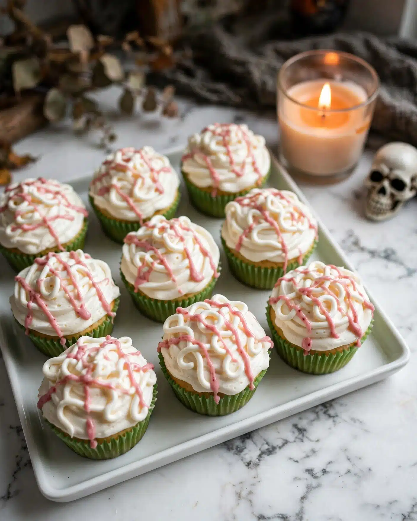 Zombie Brain Halloween Cupcakes Recipe 5 A white rectangular tray holds seven cupcakes with green wrappers. Each cupcake has a thick layer of white frosting on top, shaped to look like brain folds with squiggly lines covering the surface. Bright red syrup is drizzled over the frosting and pools around the base of each cupcake on the tray, creating a dripping effect. In the background, a lit candle in a glass holder casts a warm orange glow next to a small skull decoration. The scene sits on a white marbled surface photo taken with an iphone --ar 4:5 --v 7