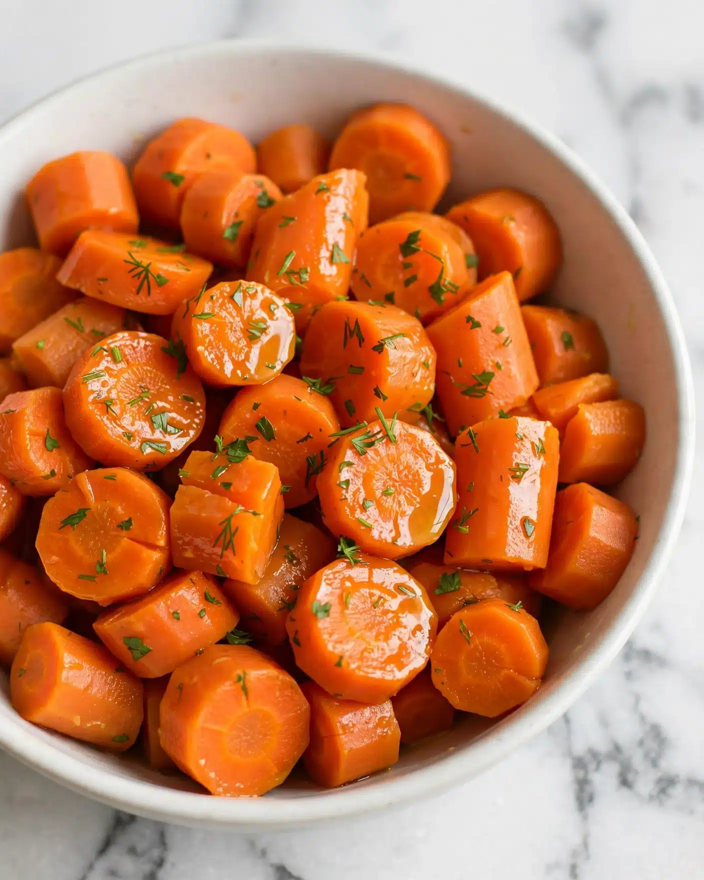 A close-up view of a white bowl filled with bright orange carrot slices that appear soft and cooked, with a slightly shiny texture. The carrot pieces are cut into uneven bite-sized rounds and chunks, and they are sprinkled with small bits of green herbs scattered evenly on top. The bowl sits on a white marbled texture background, giving a clean and fresh look. photo taken with an iphone --ar 4:5 --v 7