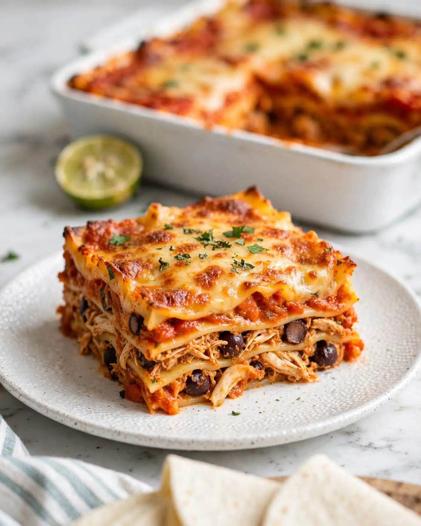 The image shows a thick, square piece of lasagna on a white textured plate, layered with orange-red tomato sauce, melted cheese in golden yellow with slight browning, and shredded pieces that appear to be chicken mixed with dark, round beans or olives. The layers of pasta sheets in pale yellow separate the dense fillings, creating a rich stacked look. The top layer is mostly covered with glossy melted cheese and a few small green herb pieces sprinkled on it. In the background, there is a white baking dish with more lasagna and a half lime, all placed on a white marbled surface. At the bottom, some white tortillas and part of a striped cloth are visible. Photo taken with an iphone --ar 4:5 --v 7
