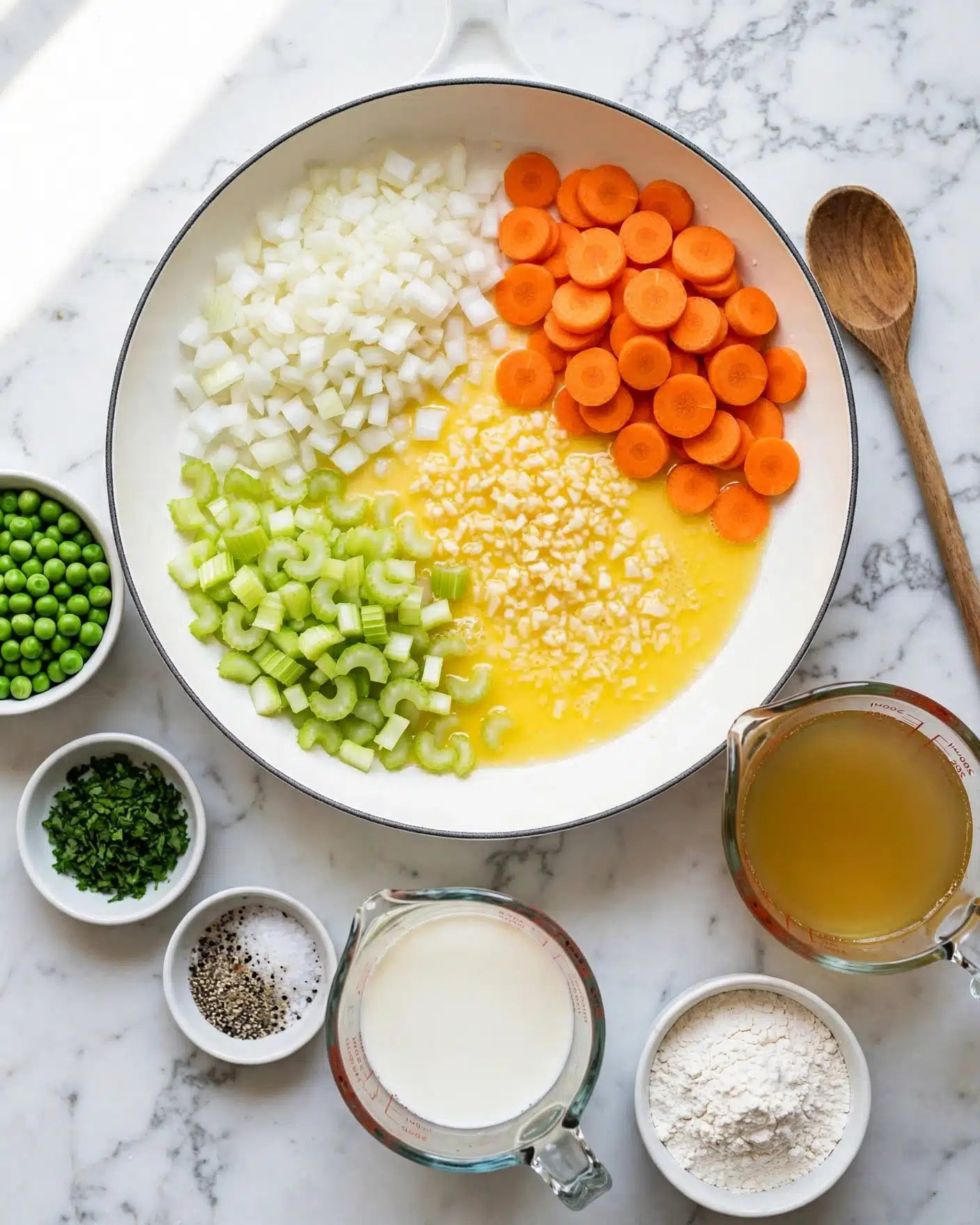 A white pan sits on a white marbled surface, filled with four sections of ingredients: diced white onions in the top left, sliced bright orange carrots on the right, chopped light green celery in the bottom left, and minced garlic pieces in the middle on a layer of melted butter or oil. Around the pan on the marbled surface are small white bowls containing green peas, fresh chopped herbs, salt and black pepper, a glass measuring cup with milk, a cup of flour, and a glass measuring cup filled with golden broth. A wooden spoon rests on the right side. Photo taken with an iphone --ar 4:5 --v 7