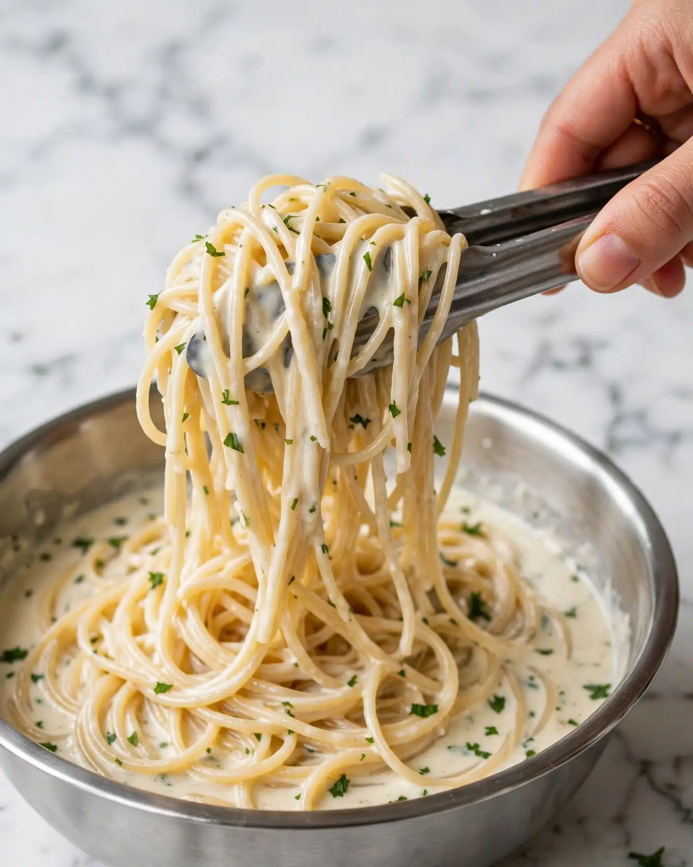 This image shows a close-up of many thin spaghetti noodles covered in a creamy, pale sauce. Small green bits of chopped herbs are scattered on top, adding a touch of color. A woman's hand is using tongs to lift a bunch of the noodles from a shiny metal bowl. The background is a white marbled texture, making the noodles stand out clearly. The noodles look soft and smooth with a slightly shiny surface. photo taken with an iphone --ar 4:5 --v 7