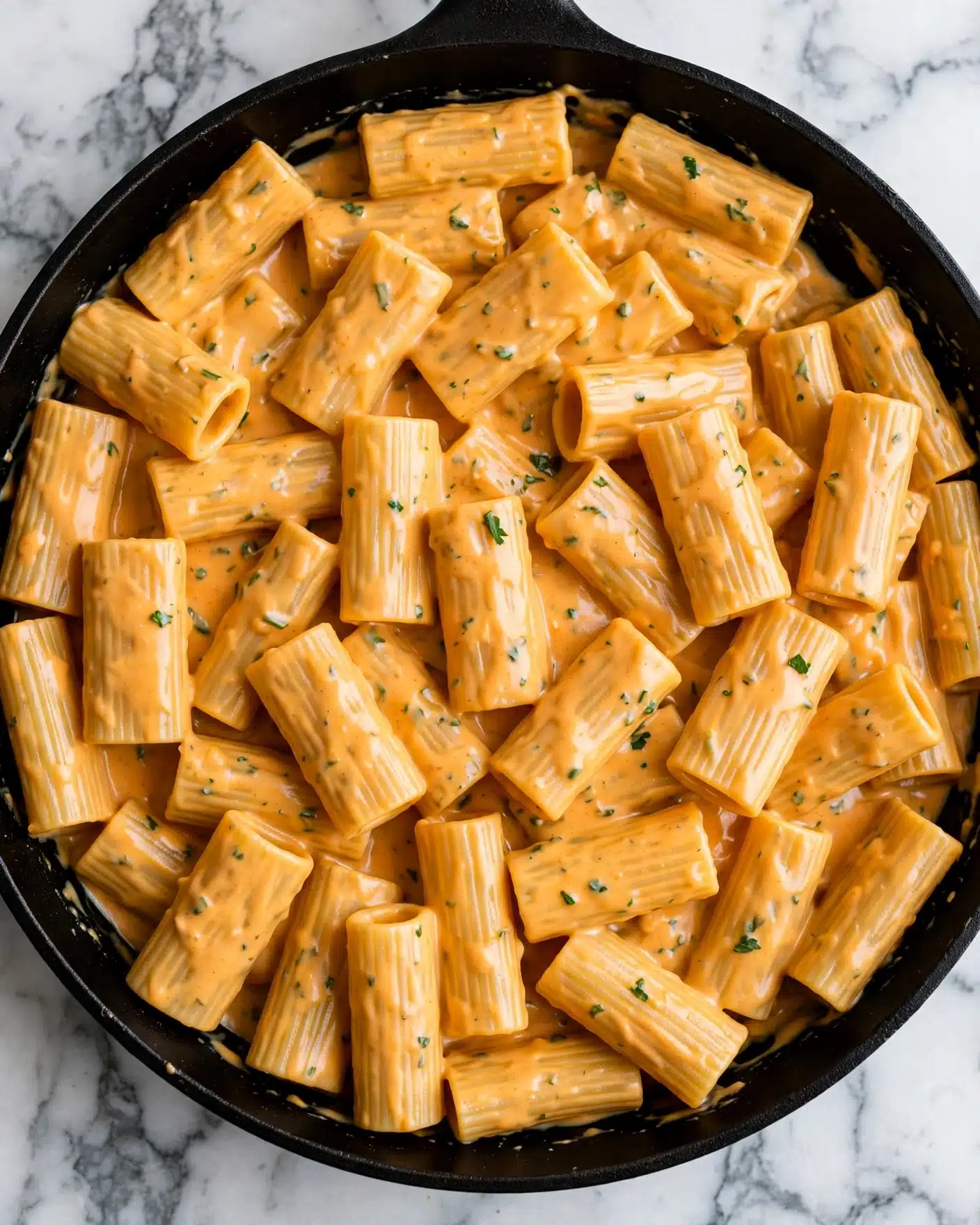 A close-up top view of a black pan filled with rigatoni pasta coated in a thick, creamy, orange cheese sauce with gentle green herb flecks throughout. The pasta pieces are large, tube-shaped, and slightly ridged, fully covered in the smooth sauce that clings to each piece. The background is a white marbled surface. photo taken with an iphone --ar 4:5 --v 7