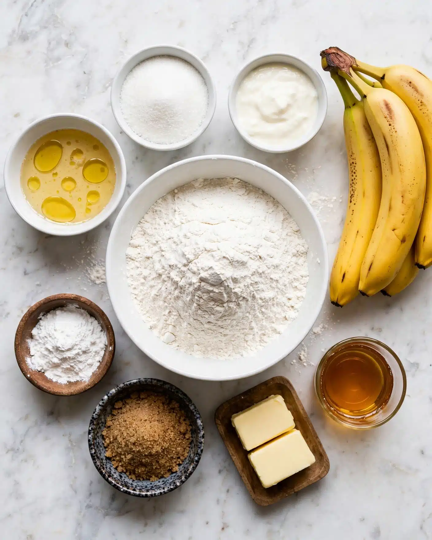 A top view of baking ingredients arranged neatly on a white marbled surface, including four ripe yellow bananas with brown spots on the right. There is a large white bowl in the center filled with white flour, surrounded by smaller white bowls holding a light golden liquid with small pools of oil, granulated white sugar, and a creamy white mixture. A small rustic bowl holds baking powder, and a wooden dish contains two blocks of pale yellow butter. Another black speckled bowl is filled with brown sugar, and a small glass cup has amber-colored syrup. The layout is clean and bright, with clear textures of each ingredient visible. Photo taken with an iphone --ar 4:5 --v 7