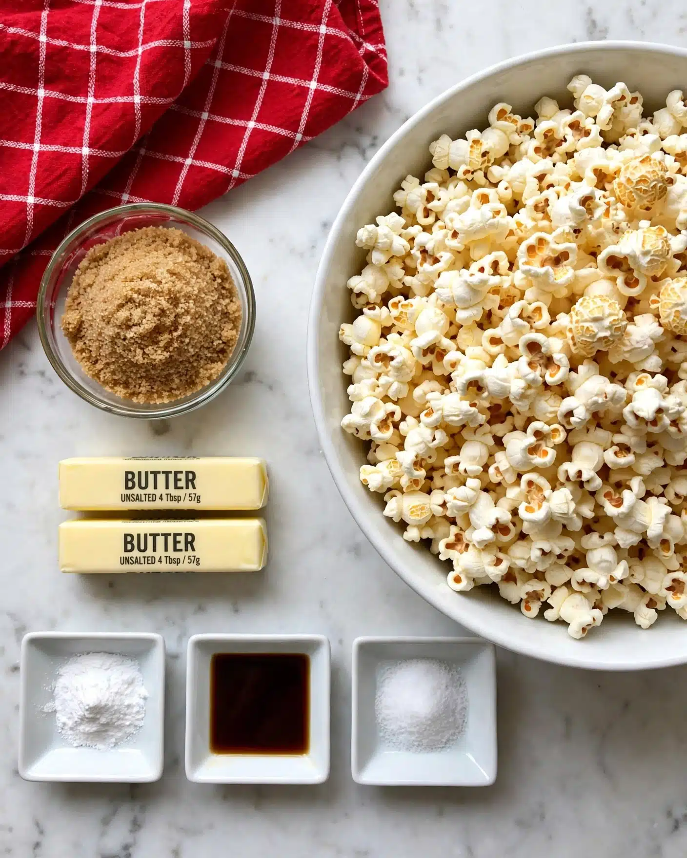 A large white bowl filled with white and light yellow popcorn, showing fluffy and slightly golden textures on top right, next to a small round glass bowl full of light brown sugar with a grainy texture on the top left; below the sugar bowl, two pale yellow sticks of butter with black text sit side by side, while beneath the butter are three small square white dishes arranged horizontally, containing white baking soda powder, dark brown vanilla extract liquid, and fine white salt respectively, all placed on a white marbled surface with a red cloth with white grid lines in the upper left corner photo taken with an iphone --ar 4:5 --v 7