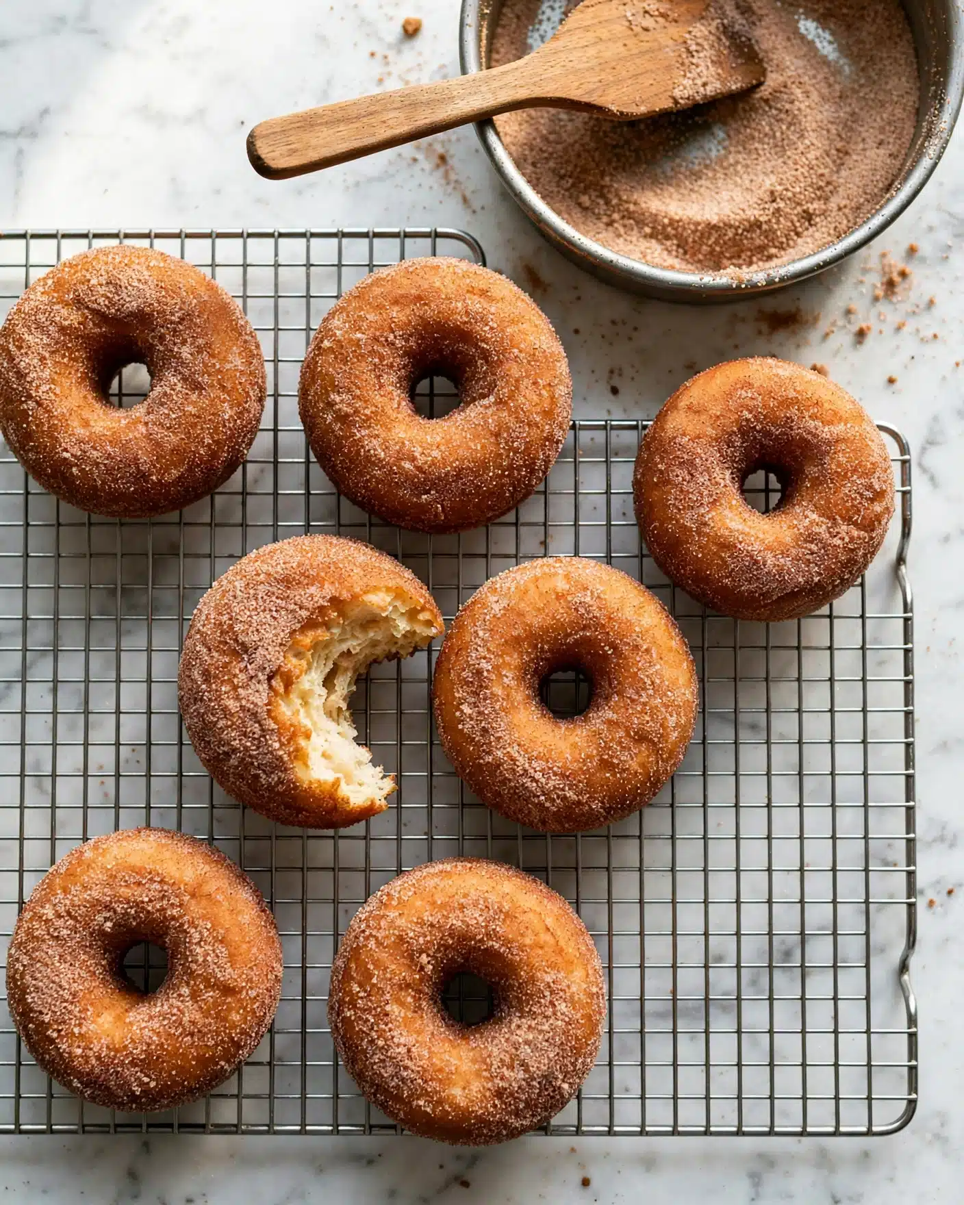 Cinnamon Sugar Doughnuts Recipe 5 Five cinnamon sugar donuts are placed on a black wire cooling rack. The donuts have a golden brown color with a grainy cinnamon sugar coating. One donut near the center has a bite taken out of it, showing a soft, yellow inside. The cooling rack sits on a white marbled surface with some scattered cinnamon sugar crumbs around it. photo taken with an iphone --ar 4:5 --v 7